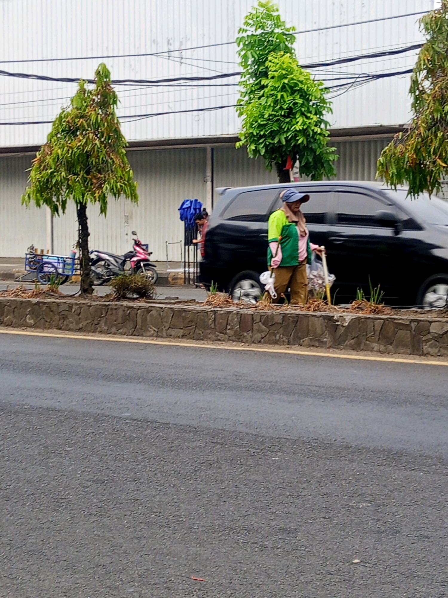 A woman wearing a blue cap, a green and pink shirt, and brown pants walks beside a black van on a street in Lubuklinggau, Indonesia. She carries a white bag and a tool with a yellow handle. A pink motorcycle and a blue cart are parked nearby. The scene is framed by a stone wall and trees, with a building in the background. The road is wet, suggesting recent rain.