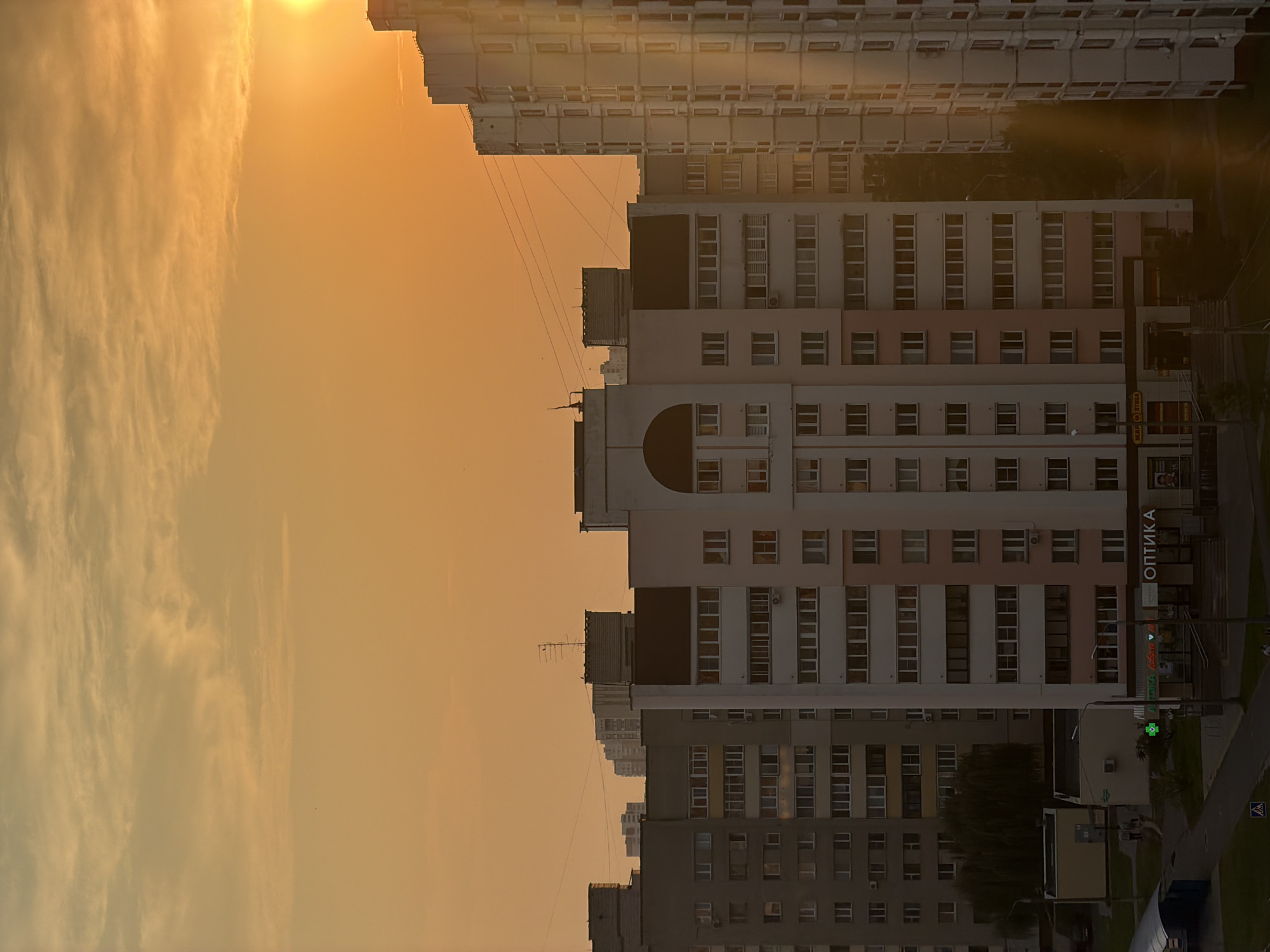 The image captures an urban landscape at sunset in Homyel, Belarus. A series of multi-story residential buildings dominate the scene, with their windows reflecting the warm, orange hues of the sky. The sky itself is filled with wispy clouds, illuminated by the setting sun which is visible on the right side of the frame. The buildings exhibit varying architectural styles and colors, from light pink and white to grey. Some buildings have visible air conditioning units and antennas. In the foreground, a street level view reveals shopfronts, including one with the Cyrillic sign "ОПТИКА," suggesting an optical store. A green cross symbol indicates a pharmacy. Several streetlights are present, and a few indistinct figures are seen walking on the pavement. The overall impression is that of a typical evening scene in a residential district.