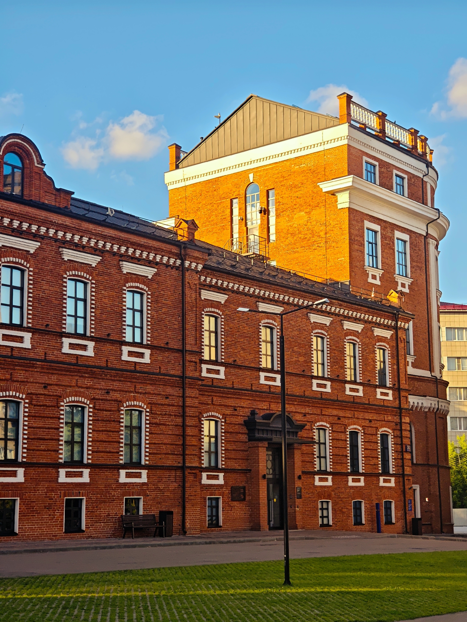 This image depicts a historic red brick building in Moscow, Russia, bathed in the warm glow of what appears to be late afternoon or early evening sunlight. The building features a prominent tower with a curved facade and a white decorative railing at the top. The main section of the building is long and rectangular, with multiple stories and numerous windows. These windows are tall and narrow, framed with white decorative elements, and have dark panes that reflect the sky and surrounding light.
The architecture is characterized by its intricate brickwork and white ornamental details, particularly along the rooflines and around the windows. The roof is a mix of dark tile and a steeply pitched metal section on the tower.
In front of the building, there is a paved walkway, a patch of green grass with a textured surface, and a street. A black lamppost stands near the entrance of the building, and a wooden bench and trash receptacle are visible along the walkway. The sky is a clear blue with a few scattered white clouds, suggesting pleasant weather. The sunlight creates strong shadows and highlights on the building's facade, indicating the time of day. A modern yellow-colored building with numerous windows is partially visible to the right of the historic structure, providing a contrast in architectural styles and suggesting an urban setting. No people are visible in the image, and no discernible text is legible on the building or its surroundings.