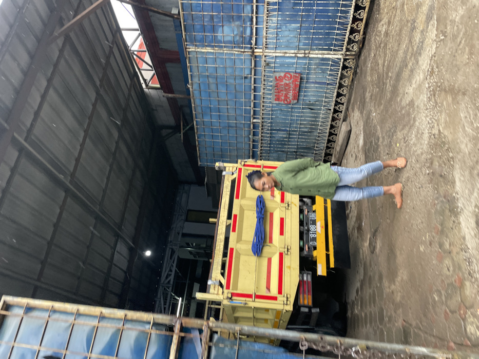 The image shows a young woman standing in front of a large, yellow dump truck. The truck has a license plate that reads "D 8188 UH". The woman is wearing a green shirt and blue jeans. She is standing in a dirt lot, looking at the camera. Behind the truck is a blue metal gate with a red sign on it. The sign is in Indonesian, and it reads "Dilarang Membuang Sampah". The sign is written in red marker and is attached to the gate. The woman is standing in a fenced-in area. There is a blue, corrugated metal wall to the left of the image, which is likely part of a building. The woman is standing under an open metal roof, which appears to be a structure or shelter that covers the lot. The image is likely taken in a industrial area of Ngamprah, Indonesia, as the coordinates provided correspond to the location of a large industrial park in Ngamprah, Bandung Barat, West Java.