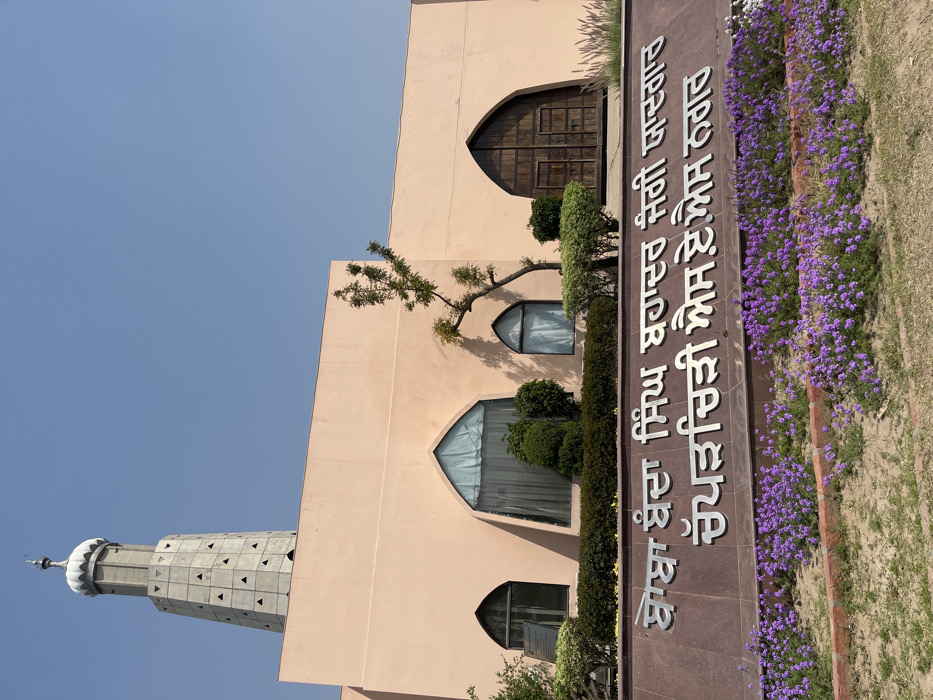 The image shows a building with a tall, thin, tower-like structure in front of it. The building is made of a light pink stucco and has several arched windows and doors. There is a sign in front of the building with words in a language other than English. There are purple flowers blooming in front of the building. The image was taken from a location in Sahibzada Ajit Singh Nagar, India, at coordinates 30.703, 76.668. The image was likely taken on a sunny day as the sky is clear blue. The image appears to be taken in an urban setting. There are no people present in the image.