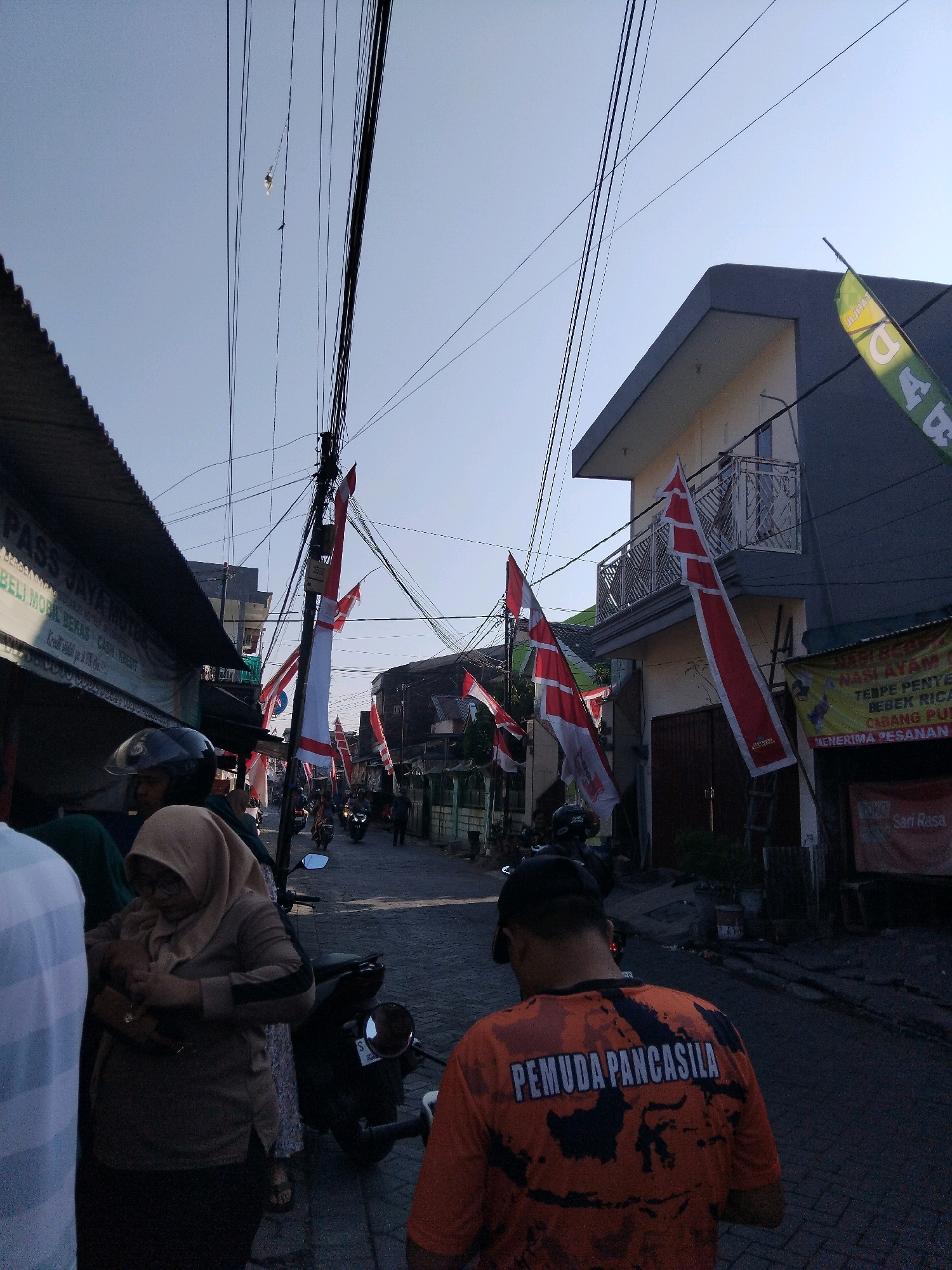 The image depicts a street scene in Wadungasri, Indonesia, possibly during a national celebration given the prominent display of Indonesian flags. The street is lined with buildings on both sides, and numerous red and white Indonesian flags are hung vertically from poles and wires. Several people are present, including individuals on motorcycles and others walking. One person in the foreground wears an orange t-shirt with the text "PEMUDA PANCASILA" and a map of Indonesia on the back. Another woman is wearing a hijab. The environment suggests a densely populated urban or semi-urban area with overhead electrical wires. Some businesses are visible along the street, indicated by signage. The overall impression is of a lively street environment with a festive atmosphere.
