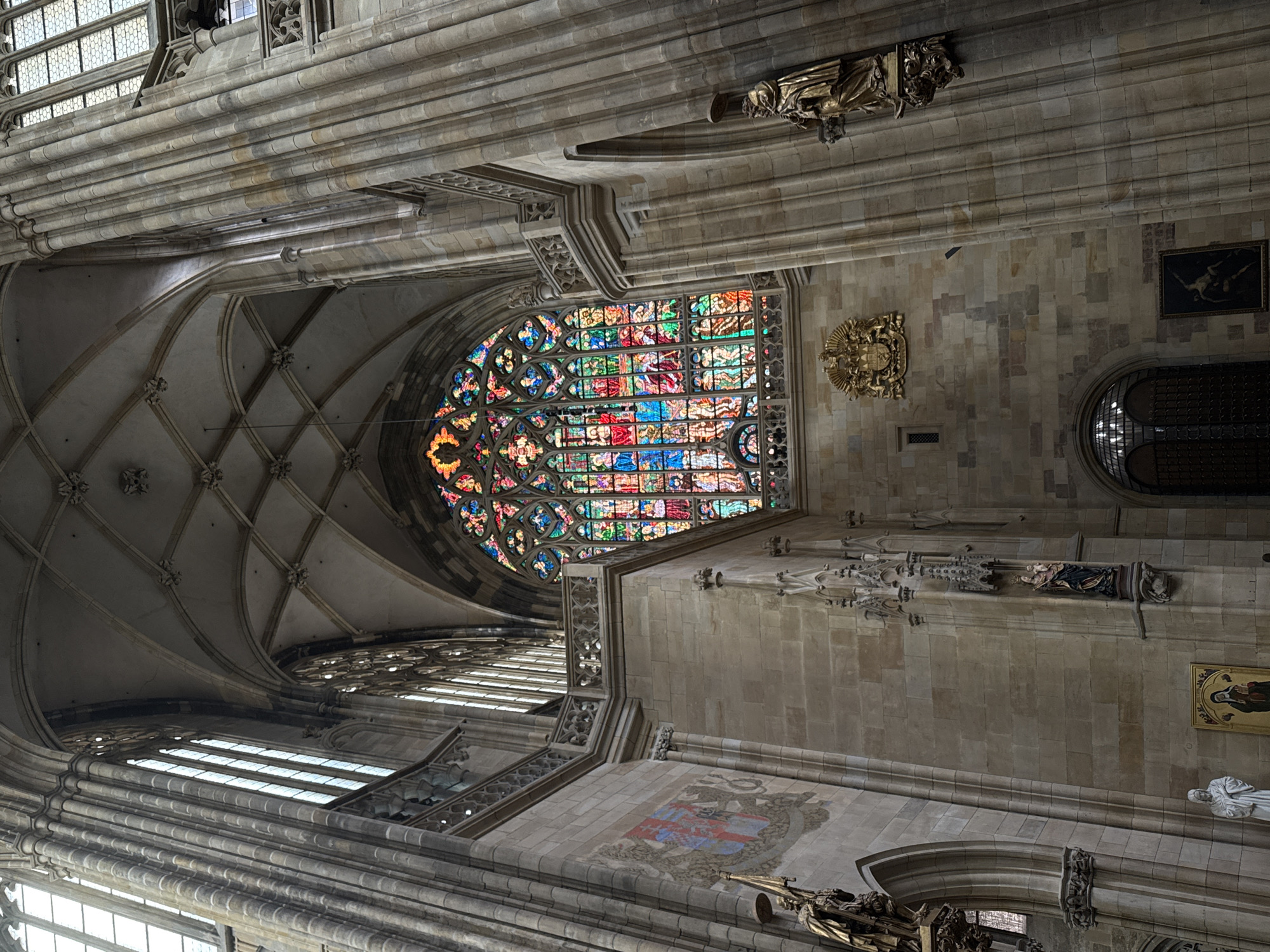 An interior shot of a Gothic cathedral shows a large, ornate stained-glass window with a circular top, featuring vibrant multicolored glass panes depicting religious scenes. The window is framed by stone architecture, including arched recesses and decorative railings. Large, fluted stone columns flank the scene, with intricate carvings and sculptures adorning the walls and niches. Sunlight streams through the window, illuminating the interior with a kaleidoscope of colors. Statues of robed figures and a coat of arms are visible on the stone walls, adding to the grandeur of the space. The ceiling exhibits a complex ribbed vaulting system with decorative bosses. The image is taken from a low angle, emphasizing the height and scale of the cathedral's interior.