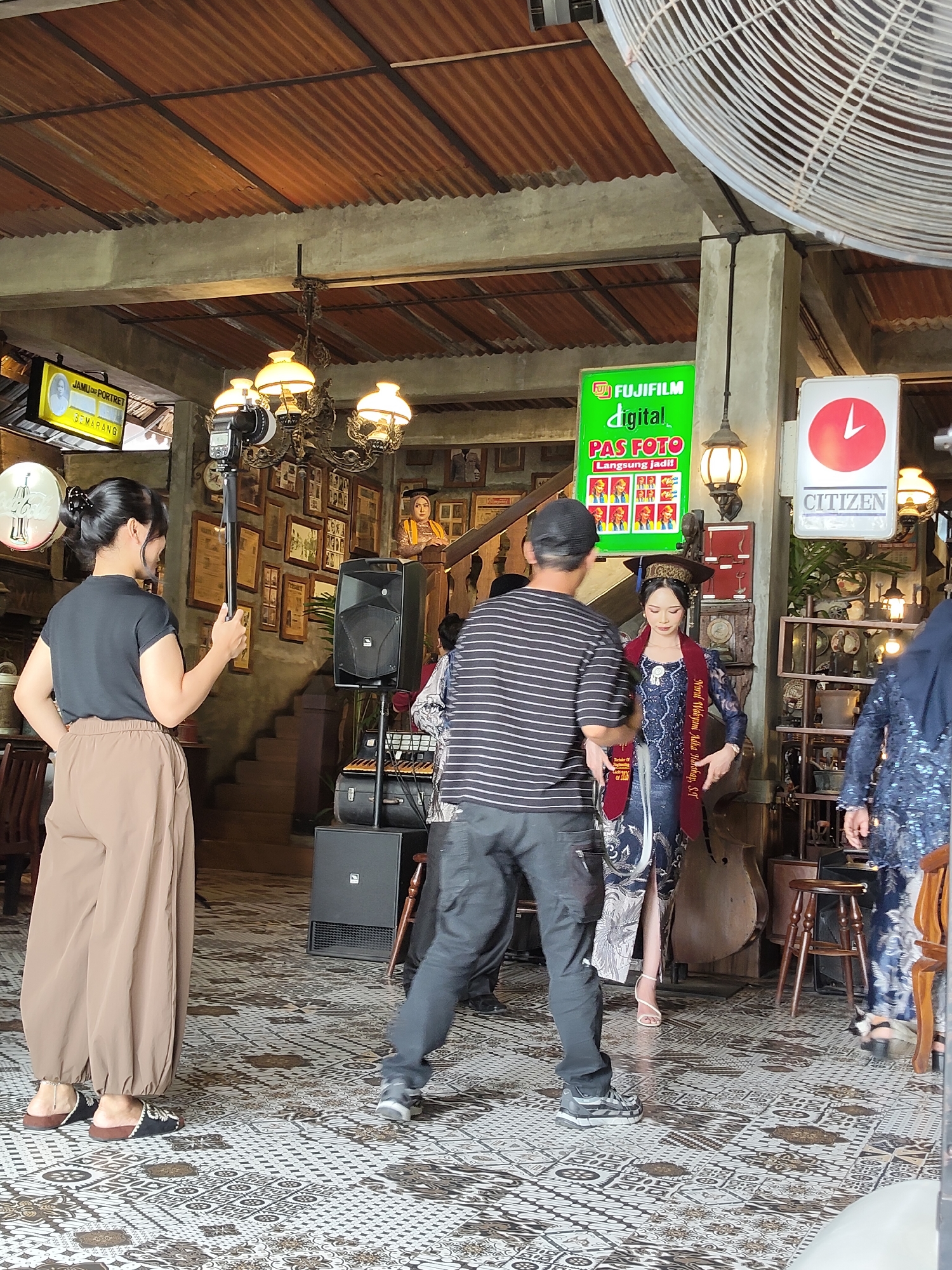 In an indoor setting with a rustic, corrugated metal ceiling and exposed concrete beams, a woman in a black t-shirt and brown pants stands facing away from the camera, holding a selfie stick with a camera attached. She appears to be recording or taking a photo of a woman in a traditional blue and gold outfit, who is facing the camera and wearing a graduation cap and sash. Behind the graduating woman, a large upright bass is visible, along with speakers and a keyboard on a stand. The room is decorated with framed photographs on the wall and vintage lamps. A green Fujifilm digital photo sign is prominently displayed on the wall, indicating a photo service. To the right, a red and white Citizen clock sign is also visible. The floor is covered in a patterned tile. The overall atmosphere suggests a celebratory event or a photo studio setup. This scene is located in the city of Medan, Indonesia.