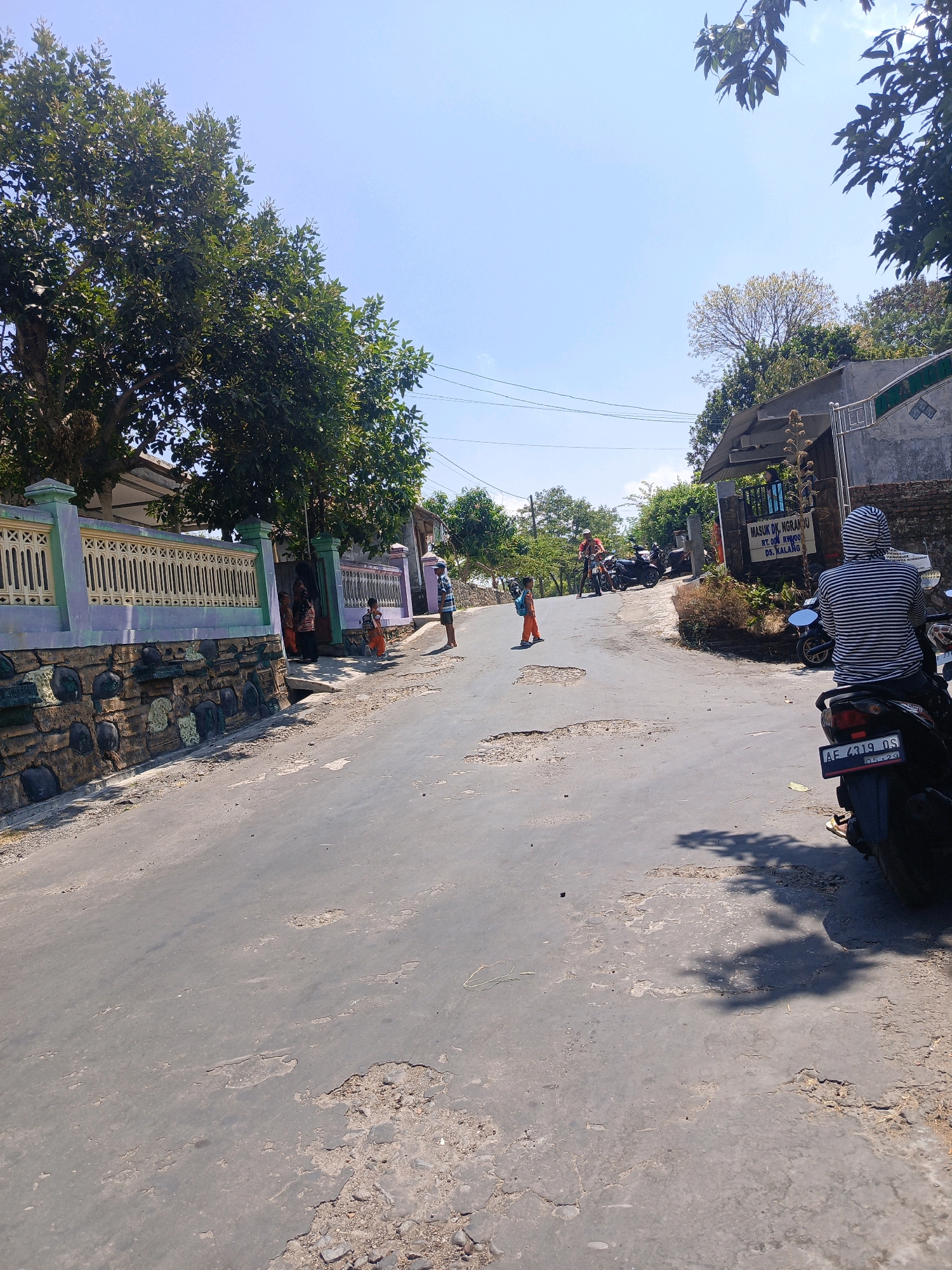 The image depicts a street scene in the village of Kalang, Indonesia. A paved road, showing signs of wear and tear with several potholes, curves uphill. On the left side of the road, a house is partially visible, featuring a decorative fence with green and purple accents and a stone wall base. Several people are gathered near the entrance of this house. Further up the road, on the right, a sign indicates "MASUK DK. NGRAHOU RT. 004 RW.009 DS. KALANG," suggesting this is an entrance to a neighborhood. Several motorcycles are parked along the road. In the foreground, a person wearing a striped hooded shirt sits on a motorcycle, facing away from the camera. Another person, a child in an orange outfit, stands on the road in the mid-ground. Trees with lush green foliage line both sides of the street, and the sky is clear blue.