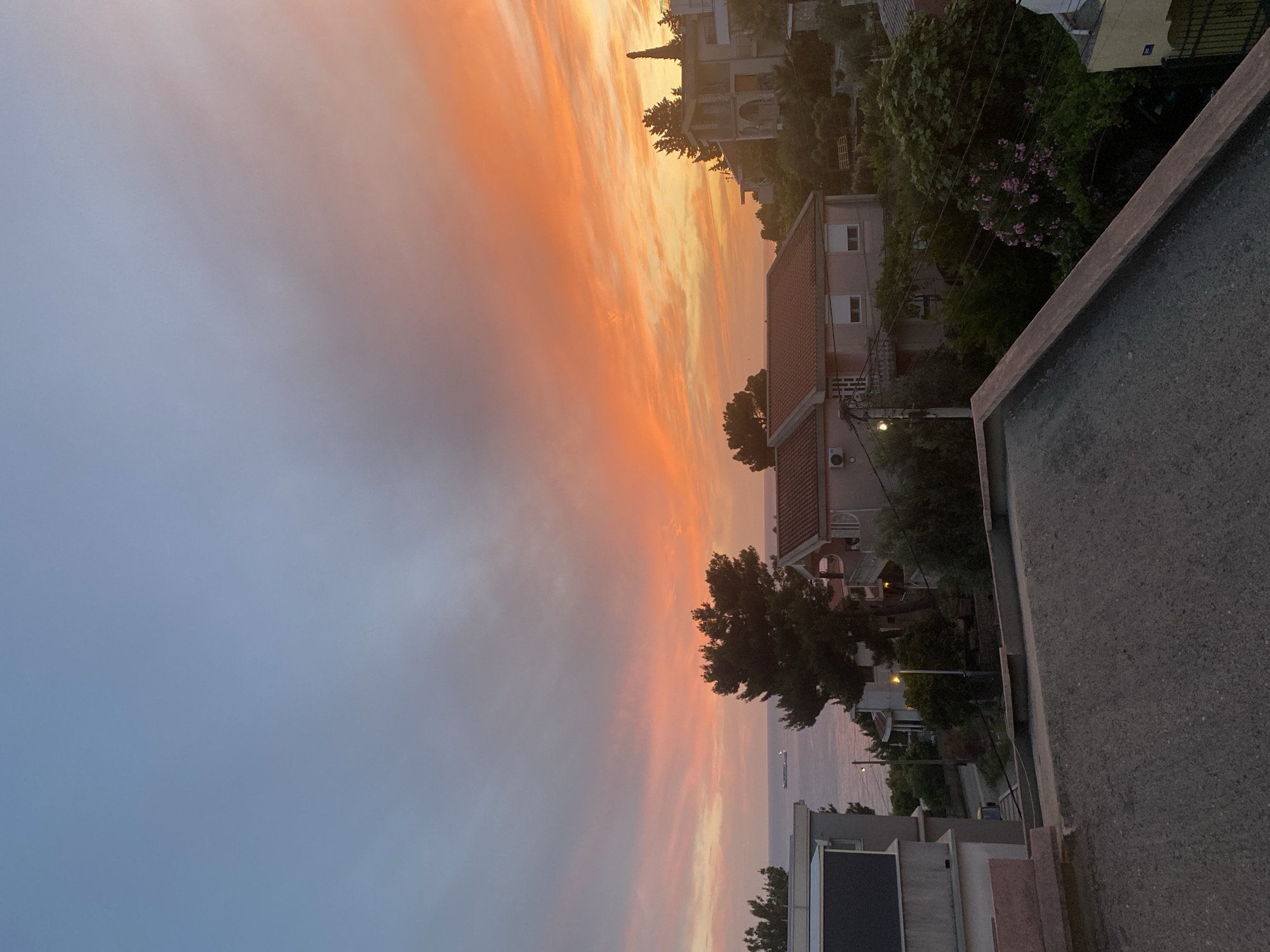 This is a photograph of a sunset over the Adriatic Sea, as viewed from an elevated position in the city of Šušanj, Montenegro. The sky is a vibrant display of orange, pink, and blue hues, with scattered clouds. In the foreground, there is a concrete rooftop with a pink-edged border. Beyond the rooftop, several residential buildings are visible, characterized by terracotta tiled roofs and light-colored walls. The architecture suggests a Mediterranean style.
One prominent building in the mid-ground has a light pink facade with white-framed windows. A large evergreen tree with dense foliage stands to the left of this building. Further to the right, another building with a more complex facade and balconies is partially obscured by trees. The sea is visible in the background, with a large ship sailing on its surface. Lush green trees and vegetation cover the hilly terrain surrounding the buildings, indicative of a coastal environment. The overall impression is a tranquil evening scene in a residential area overlooking the sea.