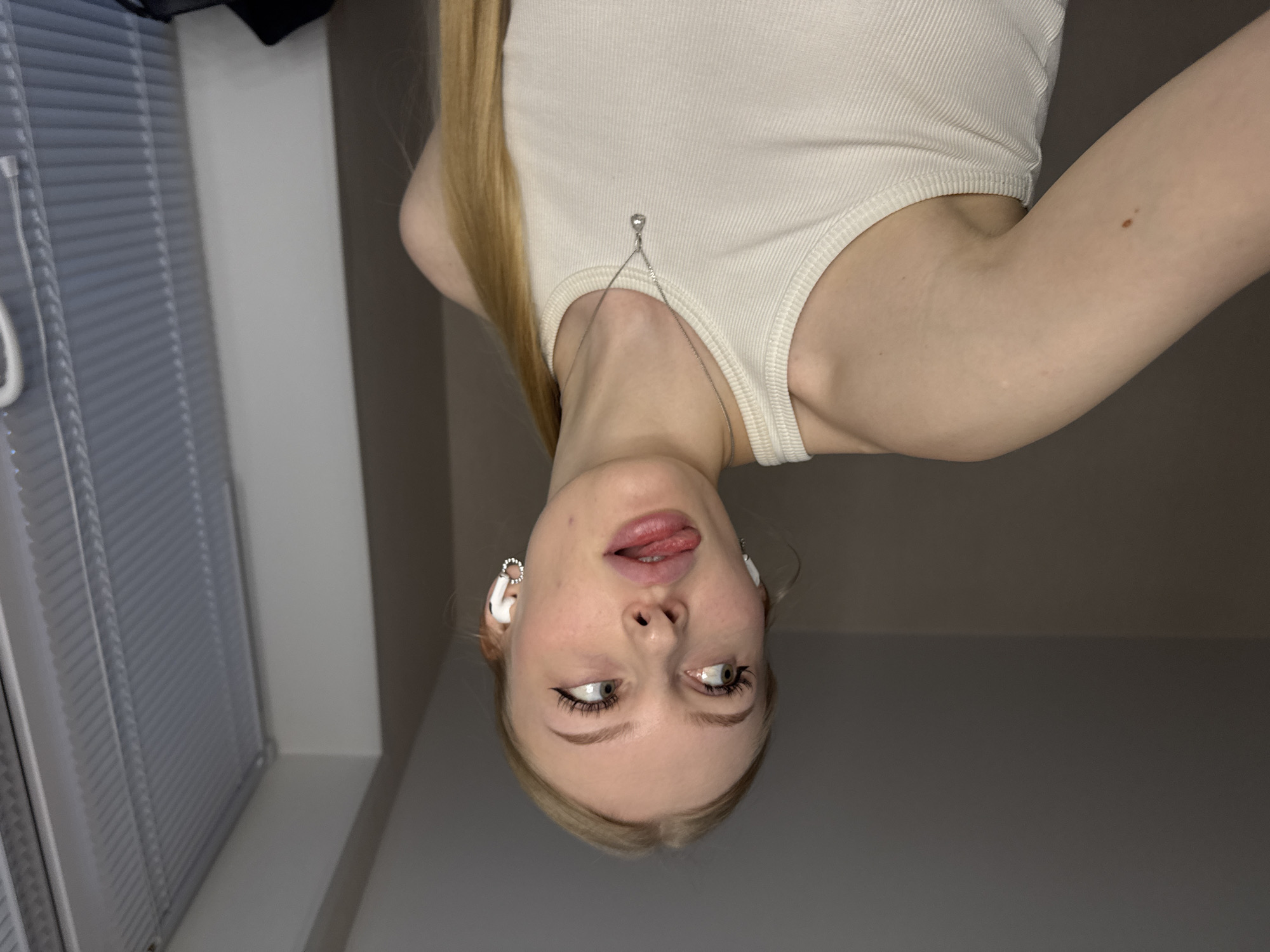 A woman is depicted upside down, wearing a white tank top with a silver chain detail at the neckline. She has blonde hair and is wearing white earbuds. Her tongue is slightly protruding, and her eyes are averted. A window with blinds is visible on the left side of the frame, and a plain wall is in the background. The location in the city of Perm, Russia, is not discernible from the visual information provided.