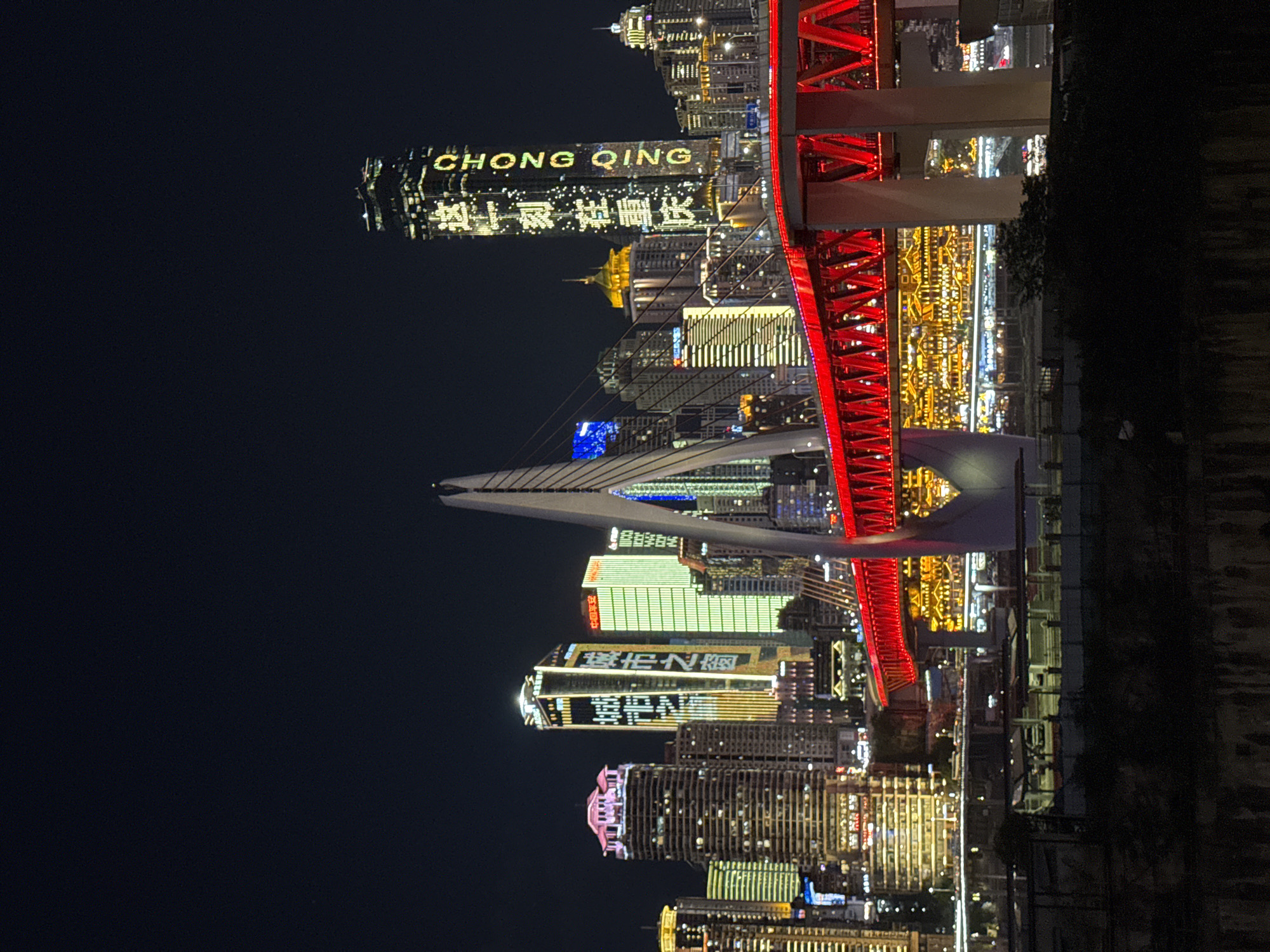 The image shows a nighttime cityscape with numerous illuminated skyscrapers and a prominent red suspension bridge. The bridge, with its red truss structure and illuminated lower deck, spans across a body of water, reflecting the bright lights of the city. A tall, slender white pylon supports one side of the bridge. Several skyscrapers of varying heights and architectural styles are visible in the background, many adorned with colorful lights. One skyscraper on the right displays illuminated text in both Chinese characters and English, reading "CHONGQING". The scene is set against a dark night sky, emphasizing the artificial illumination of the urban environment. The ground in the foreground appears to be a dark, textured embankment or wall. Based on the visible text and architectural style, the location is likely the cityscape of Chongqing, specifically around the Jiangbei City Street (江北城街道) area, which is known for its modern architecture and prominent bridges. There are no people or identifiable actions depicted in the image.
