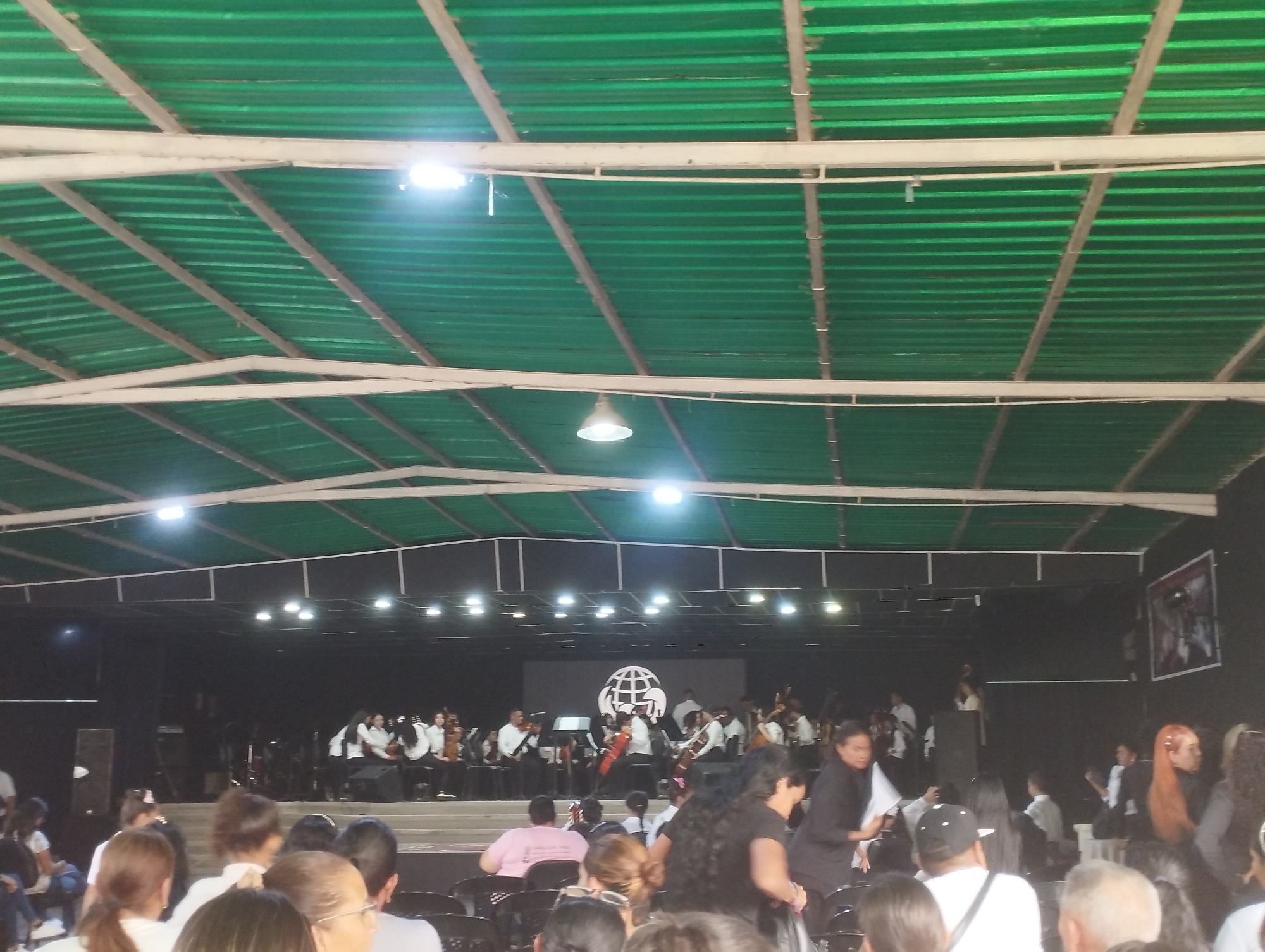 The image shows an indoor event in Tinaquillo, Venezuela. A crowd is seated in rows facing a stage where an orchestra is performing. The performers are wearing black and white attire. Behind the orchestra is a large screen displaying a logo. The venue has a green corrugated metal roof supported by white metal beams. There are lights hanging from the ceiling. Some people in the audience are walking around.