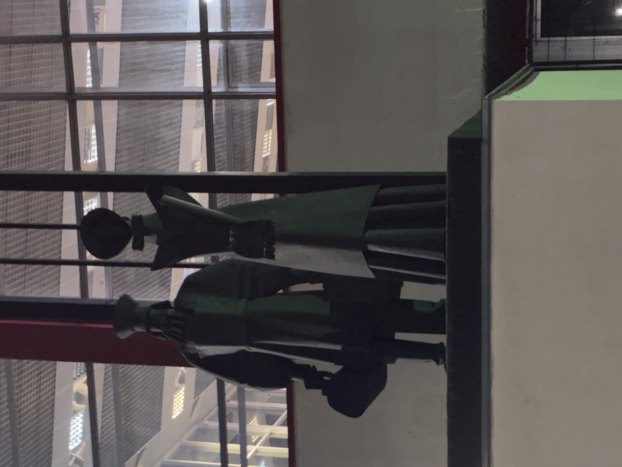 The image shows a statue of two figures, likely a couple, standing in front of a building with a grid-like window structure. The figure on the left appears to be male, wearing a hat and holding a bag. The figure on the right is taller and seems to be wearing a dress. The statue is dark in color and appears to be made of metal. The background consists of a building with large windows and what looks like an interior with lighting. The scene suggests an outdoor setting in a city. The image is taken at night or in low light conditions, with some artificial lighting visible in the background. The overall impression is that of a public art installation. This specific statue is located in Lipetsk, Russia, at the "Sculpture of the Family" on Sobornaya Square.