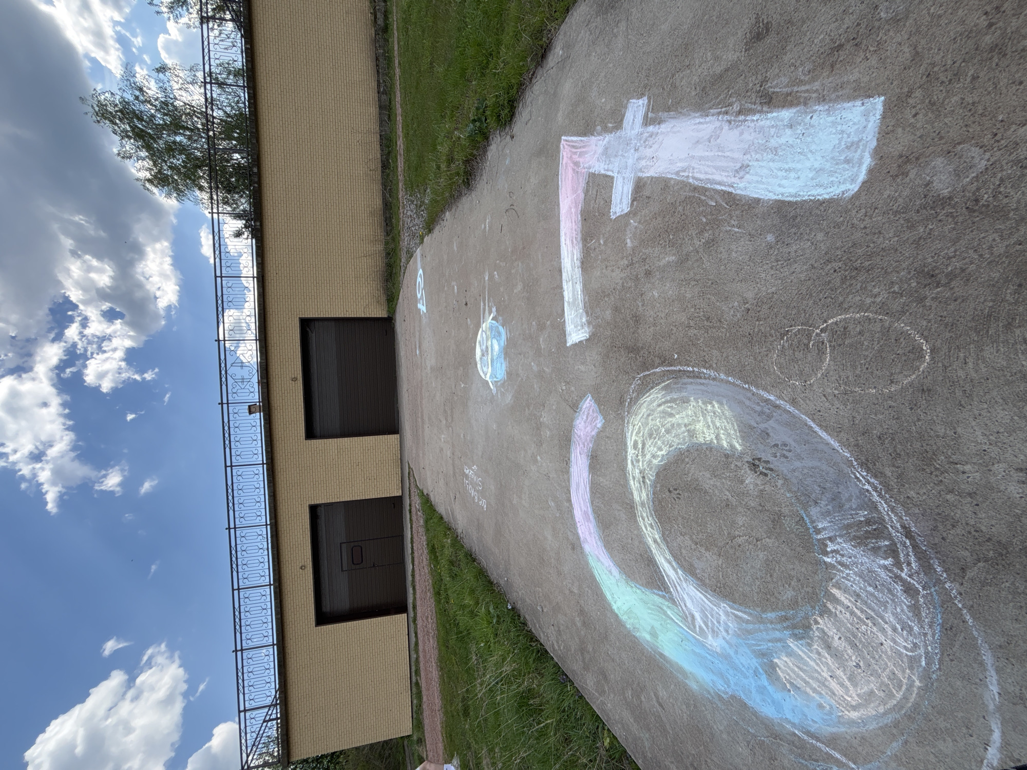 A ground-level perspective shows a concrete path with chalk drawings on it. The drawings include the numerals "6," "7," and smaller circular shapes. To the left of the path is a two-story building with a yellow brick facade, dark-colored garage doors, and a decorative black railing along the top of the building. Adjacent to the path is grass. The sky is visible with clouds. The location appears to be outdoors, possibly in Овсянка, Russia.