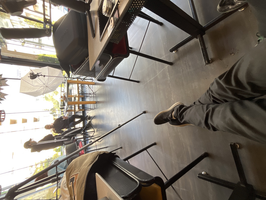 The image depicts an indoor scene within a modern establishment in Kafr Sousa Municipality, Syria, likely a cafe or restaurant, where a media production is underway. The perspective is from a seated individual, whose legs, clad in black trousers and black sneakers, extend into the foreground. To the left, a dark-colored jacket or bag rests on a table or chair. To the right, a black-framed table with a light-colored top features a recessed area containing small metallic objects, possibly production tools. Black tripod legs are visible on the dark tiled floor between the viewer and the central activity.
In the midground, illuminated by natural light entering through large windows, a man in a dark hoodie and cap stands behind a camera setup on a tripod. He faces another man seated at a light-colored wooden table. The seated man, dressed in dark attire, gestures with his hand near his face, with food items visible on the table before him. To his right, a large white photographic umbrella (softbox) is positioned, indicating artificial lighting for the production. Additional chairs and tables are arranged around the space, and a large black equipment case rests on a chair on the right side of the frame. Through the expansive windows in the background, outdoor trees and multi-story buildings are visible under daylight conditions. Decorative banners, one of which appears to read "SYRIA," hang from the ceiling.