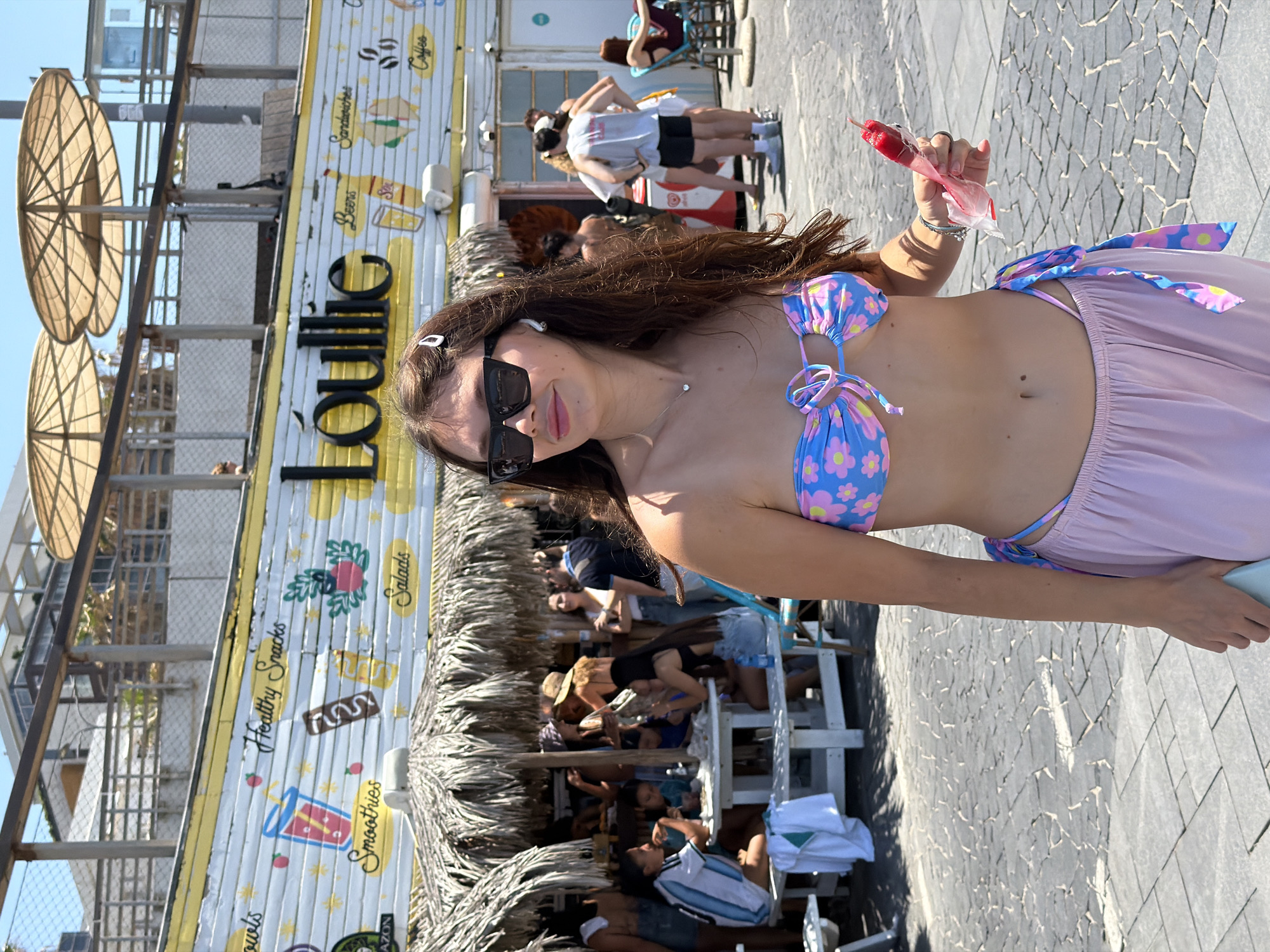 A young woman is holding a partially eaten popsicle, standing next to a beach-side restaurant named "Loullie" in Tel-Aviv, Israel. The restaurant's exterior is decorated with images of smoothies, salads, sandwiches, and other food items. Behind the woman and the restaurant, patrons are seated at tables under a thatched roof and sun umbrellas. Other people are visible walking in the background.