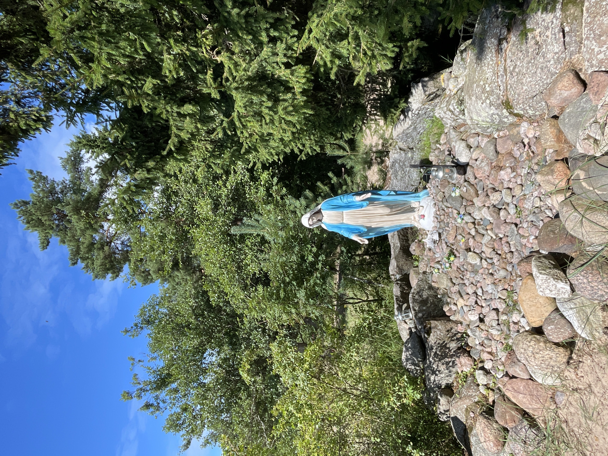 A statue of the Virgin Mary stands amidst a rocky, natural setting, likely in Zelenaya Roshcha, Russia. The statue is dressed in a blue robe over a cream-colored gown, with her head bowed and hands open. It is positioned on a mound of gravel and rocks, surrounded by larger stones. A small, lantern-like object and some flowers are placed near the statue. Lush green trees, including pine trees, form a dense backdrop, with a bright blue sky visible above. The ground around the rocks appears sandy with some grass. The scene conveys a sense of serene outdoor devotion.