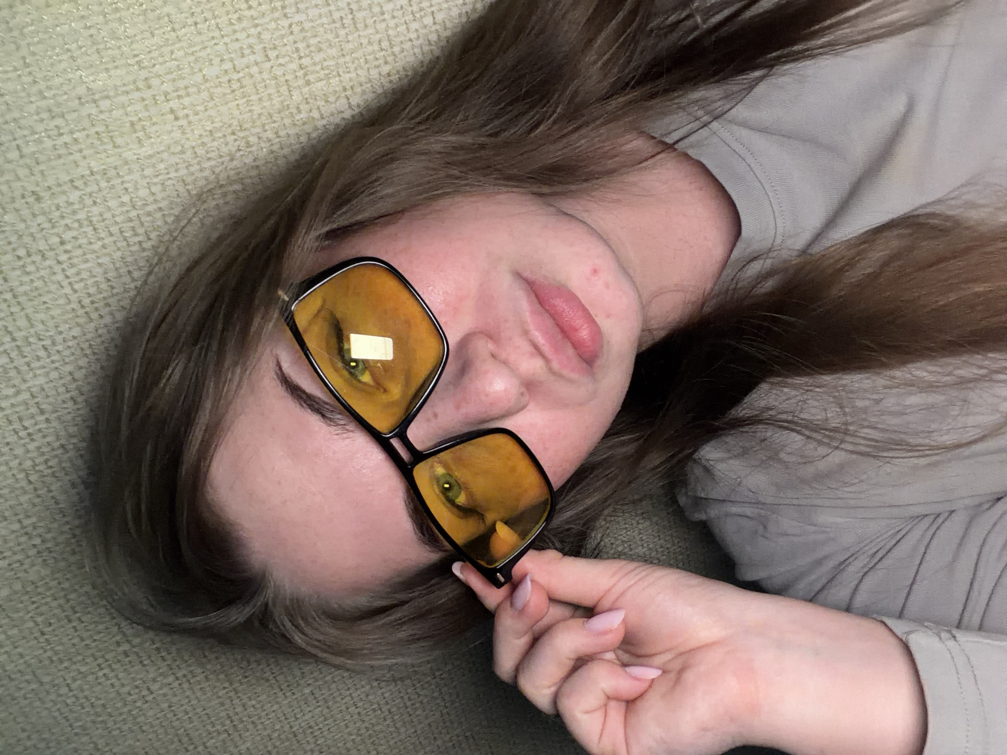 A woman with long brown hair lies on a textured surface, possibly a couch or bed. She wears glasses with amber-tinted lenses. Her right hand holds the corner of the glasses. She appears to be wearing a light-colored shirt or top. There is no information to suggest this photo was taken in the city of Chaikovsky, Russia.