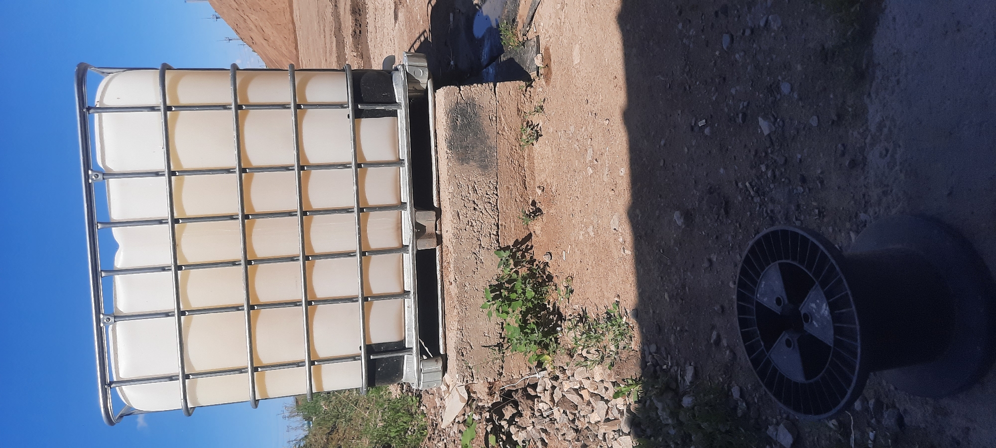 The image depicts an outdoor scene on a sunny day, with a bright blue sky and scattered clouds. In the foreground, a black plastic cable spool rests on a gravelly, sandy surface. The ground is uneven, with visible stones and debris.
Dominating the middle ground is a large, white IBC (Intermediate Bulk Container) tote. This container is encased in a silver metal cage and sits on a concrete platform. The tote is partially filled with a light-colored liquid. The concrete platform appears weathered and is surrounded by more gravel and some sparse green vegetation.
In the background, to the right, a large pile of reddish-brown earth or sand rises up. The overall environment suggests an industrial or construction site, possibly in a rural or undeveloped area. There are no people visible in the image. The location is identified as Zhasagan, Kazakhstan.