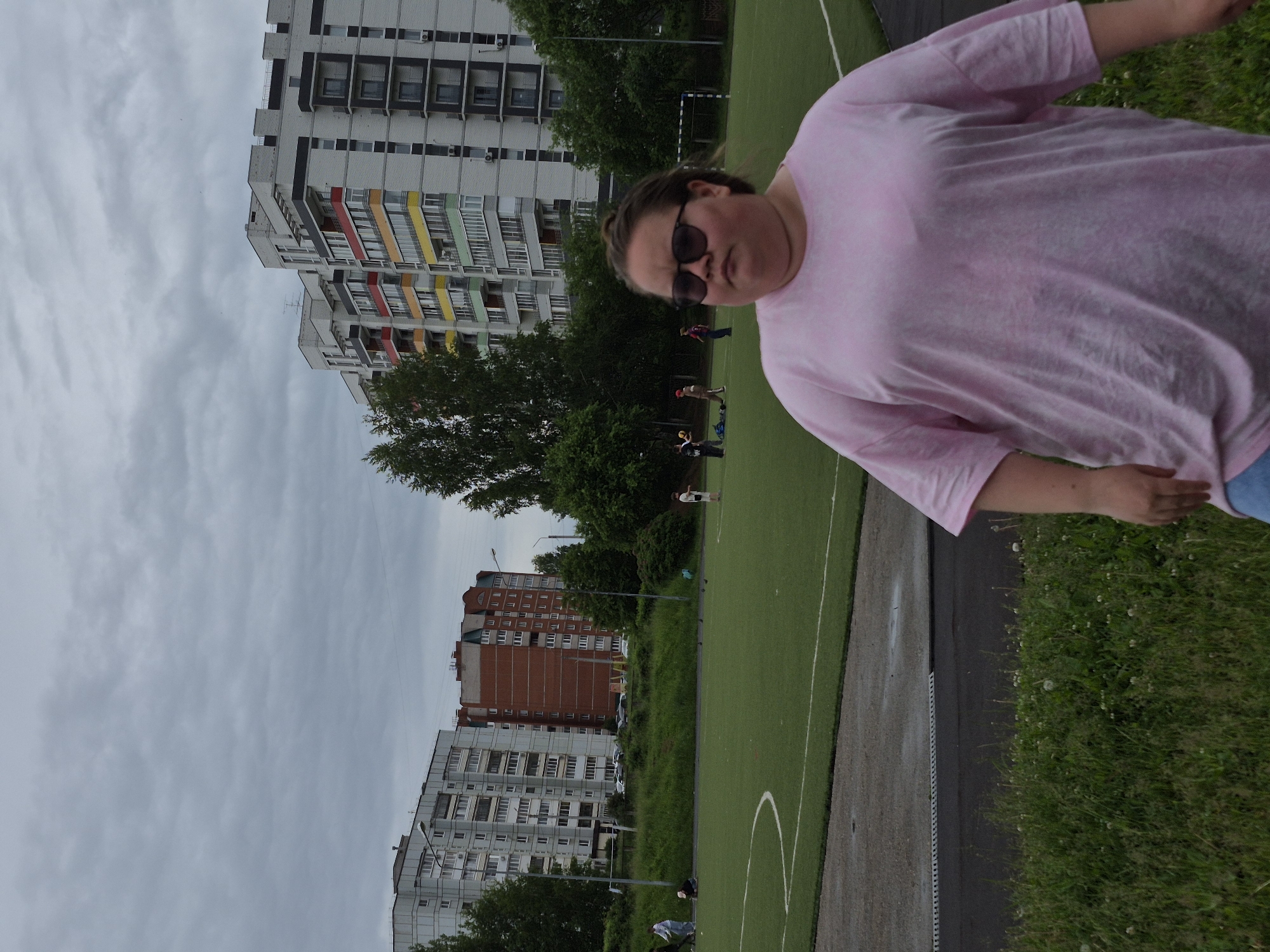 The image depicts an outdoor urban scene featuring a woman in the foreground and a sports field bordered by residential buildings. The main subject, a woman, is positioned on the right side of the frame, wearing a light pink, possibly tie-dye, t-shirt and dark sunglasses. Her expression is neutral, and her hair is pulled back.
In the mid-ground, a green sports field with white markings, resembling artificial turf for soccer, stretches across the scene. A white goal post is visible in the distance on the field. Several other individuals, appearing to be children or young adults, are sparsely distributed across the field, suggesting casual recreational activities.
The background is dominated by multi-story apartment buildings. One prominent building features a modern design with white and grey sections and balconies painted in vibrant colors like red, yellow, green, and light blue. Another, further back, is a red-brick building. Trees and lush green foliage are interspersed between the buildings and alongside the sports field.
The sky is uniformly overcast and grey, indicating a cloudy daytime setting. The architecture of the buildings and the general urban environment are consistent with residential areas in Chaikovsky, Russia. There is no visible text in the image.