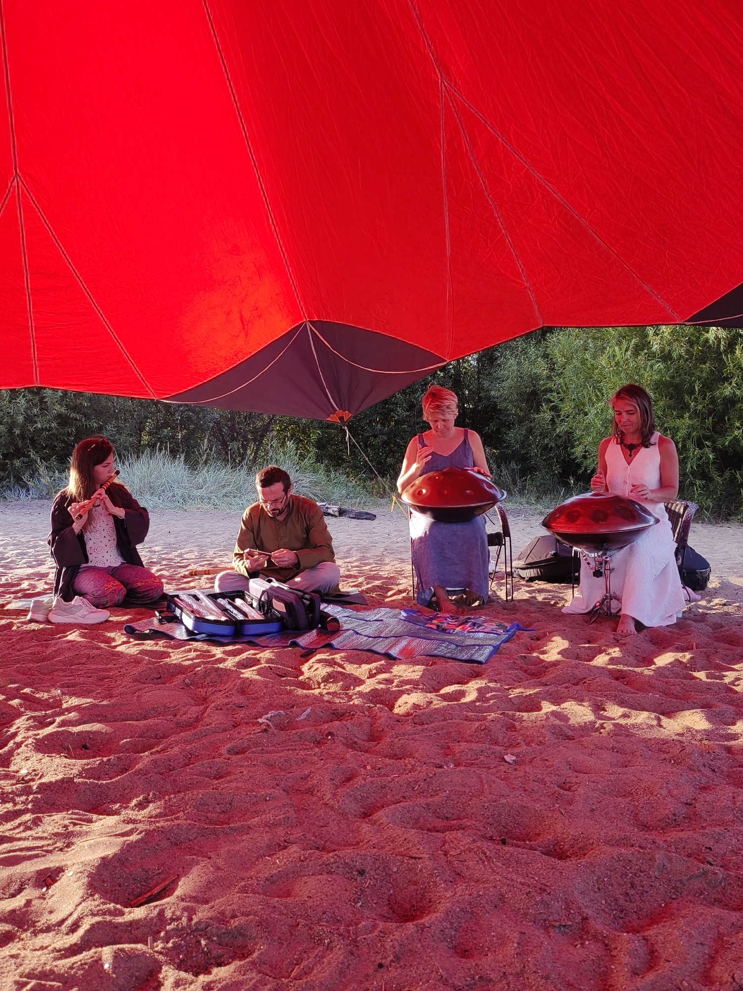 Four individuals are performing music on a sandy beach under a large, red canopy. In the foreground, a woman sits on the sand and plays a flute. To her right, a man sits on the sand and appears to be manipulating a small object. In the background, two women are seated on chairs, each playing a handpan. One woman wears a blue dress, and the other wears a white dress. The environment is a beach with sand and vegetation visible behind the performers. The image is cropped to show only a portion of the scene, with the red canopy dominating the upper part of the frame and the sand filling the lower part. The lighting suggests it is daytime, possibly late afternoon due to the warm tones. No specific Saint Petersburg landmarks are visible.