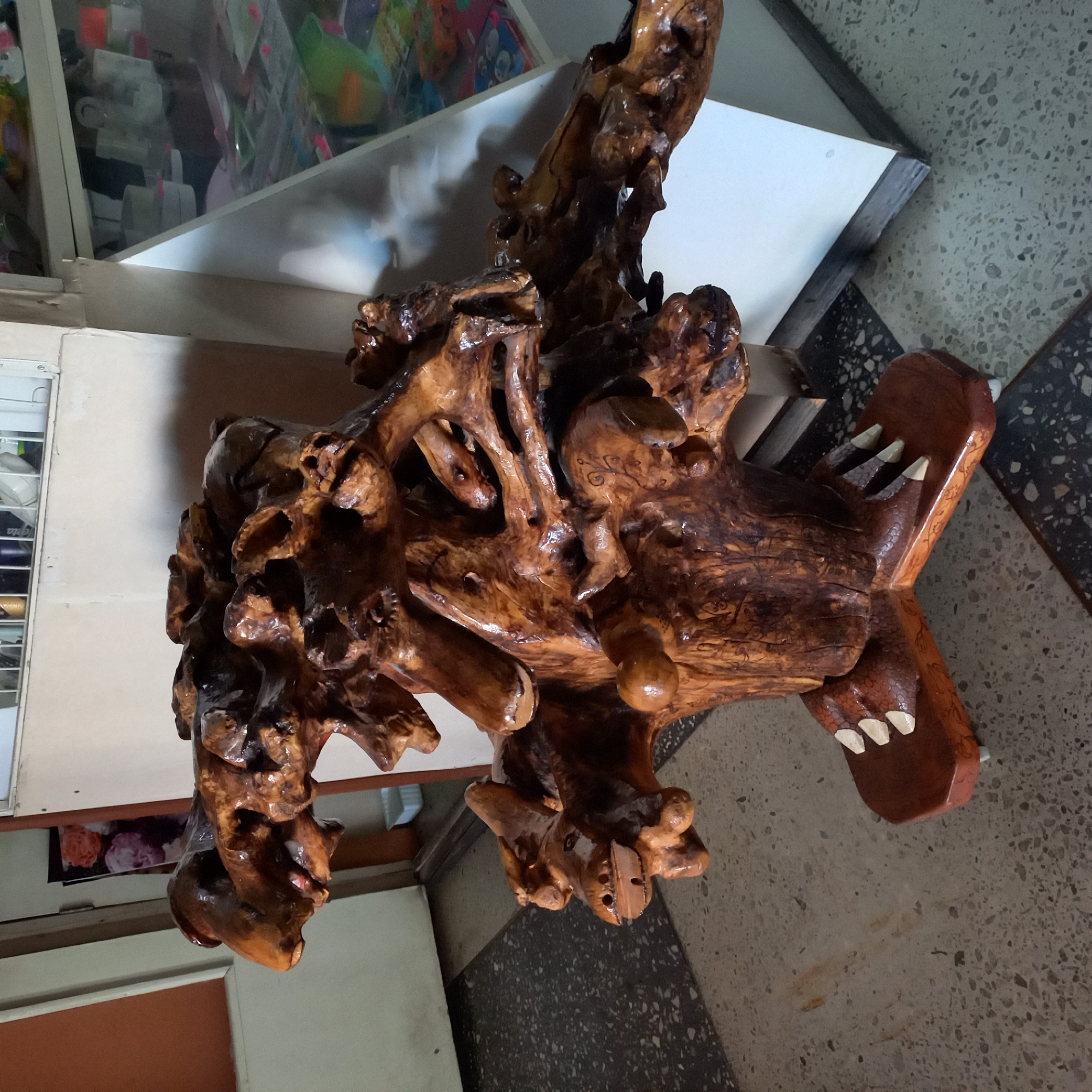 The image shows a wooden sculpture of a mythical creature, possibly a dragon or a griffin, situated indoors. The sculpture is made from a large, gnarled piece of wood, with various protruding elements that form the creature's head, limbs, and body. The wood has a polished, dark brown finish, highlighting its natural grain and texture. The creature appears to have multiple heads or faces, with open mouths and detailed carved features. It rests on a carved wooden base that resembles claws or talons, which in turn sits on a patterned tiled floor. In the background, there is a doorway with an orange-colored wall on the left, and to the right, a display case with various items inside, suggesting a shop or a public space. The overall environment appears to be dimly lit. The specific location is identified as the city of Kropyvnytskyi, Ukraine. No people are visible in the image.