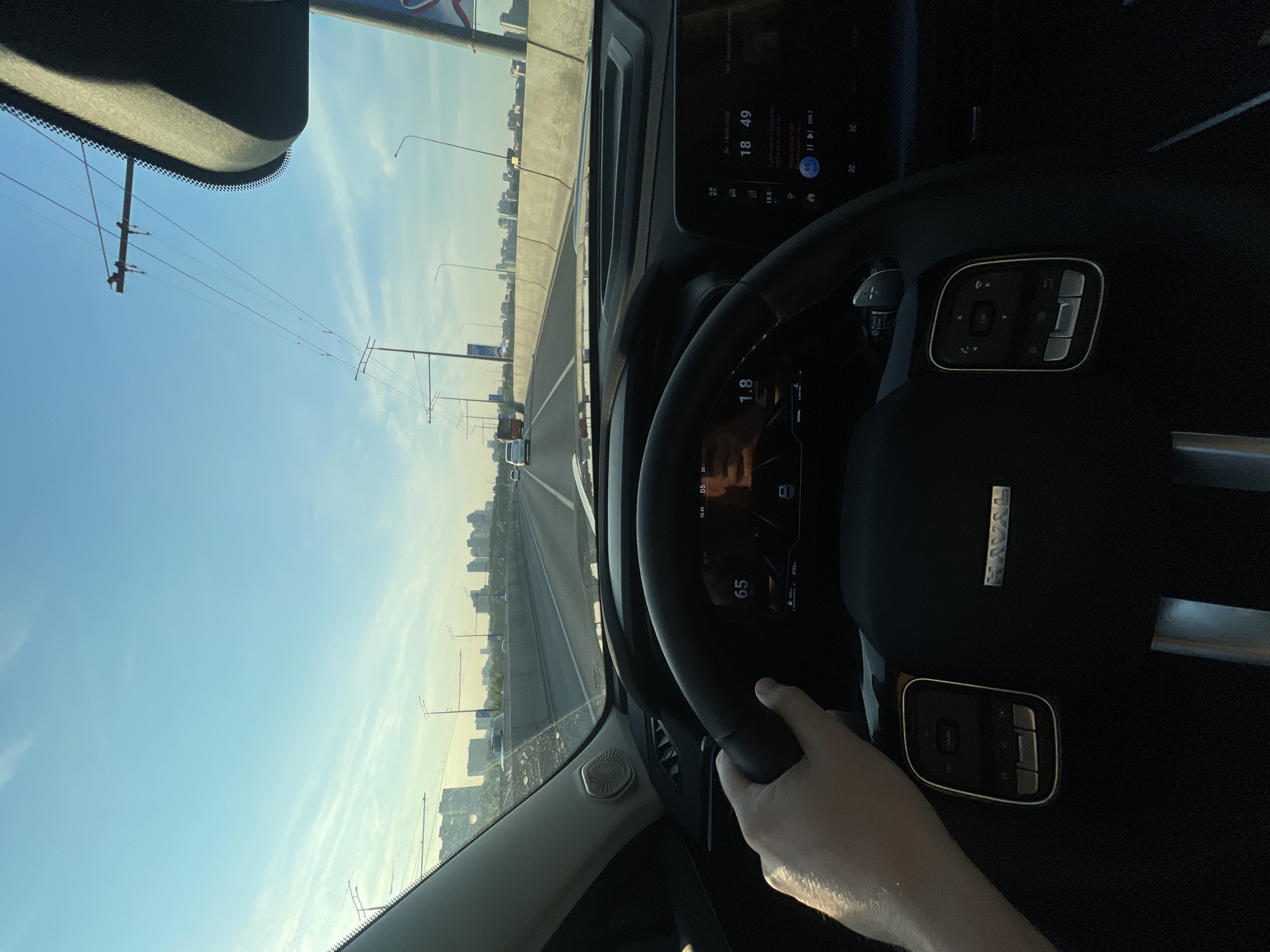 A first-person perspective captures the interior of a car driving on an elevated highway during the daytime. The driver's left hand is visible on the steering wheel, which features control buttons and the "HAVAL" logo. The dashboard displays the car's speed as 65 km/h and a digital clock showing 18:49, with the date 14/06/2020. The view through the windshield shows a highway stretching into the distance, flanked by a concrete barrier and city buildings on either side. Overhead power lines are prominent against the clear blue sky with wispy clouds. The overall scene suggests a typical commute or journey through a developed urban area.
