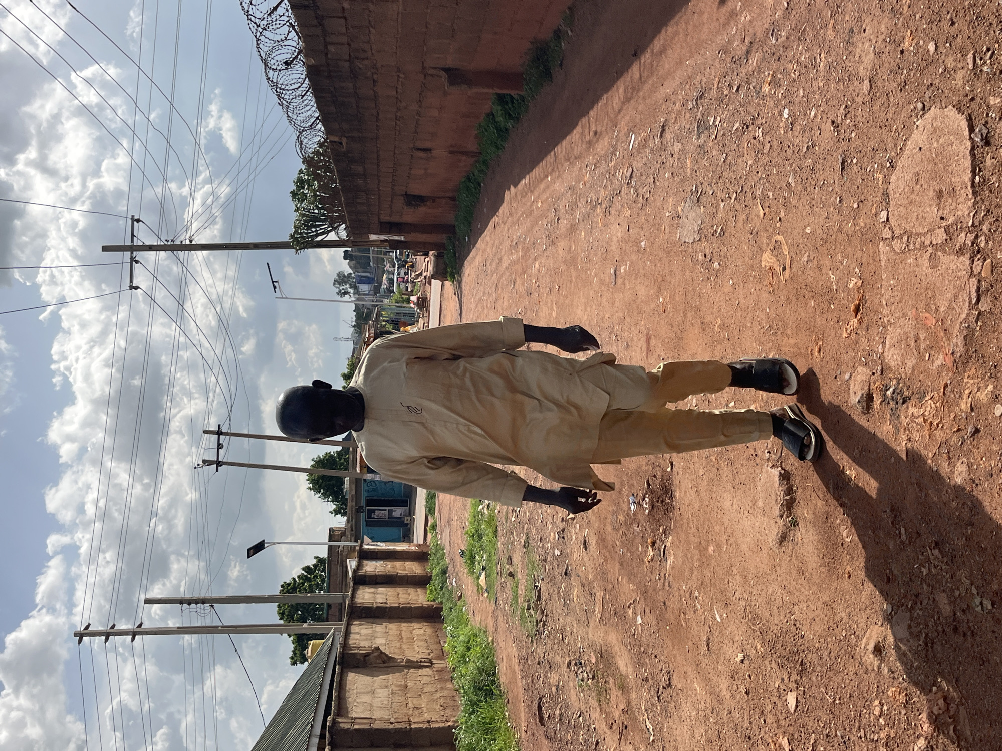 An outdoor, eye-level, medium shot captures a man walking on a dirt road under a cloudy sky in a city. The man, with his face obscured by shadow, is wearing a light-colored, long-sleeved tunic and pants, and black sandals. He is walking away from the viewer towards the center of the frame. The road is unpaved and dusty, with scattered debris. On the left, a brick wall with a corrugated metal roof runs alongside the road. On the right, a taller brick wall with barbed wire on top borders the road, extending into the background. Utility poles with numerous wires are visible in the sky and above the walls, suggesting the presence of infrastructure. In the distance, more buildings and trees can be seen. The lighting indicates it is daytime.
