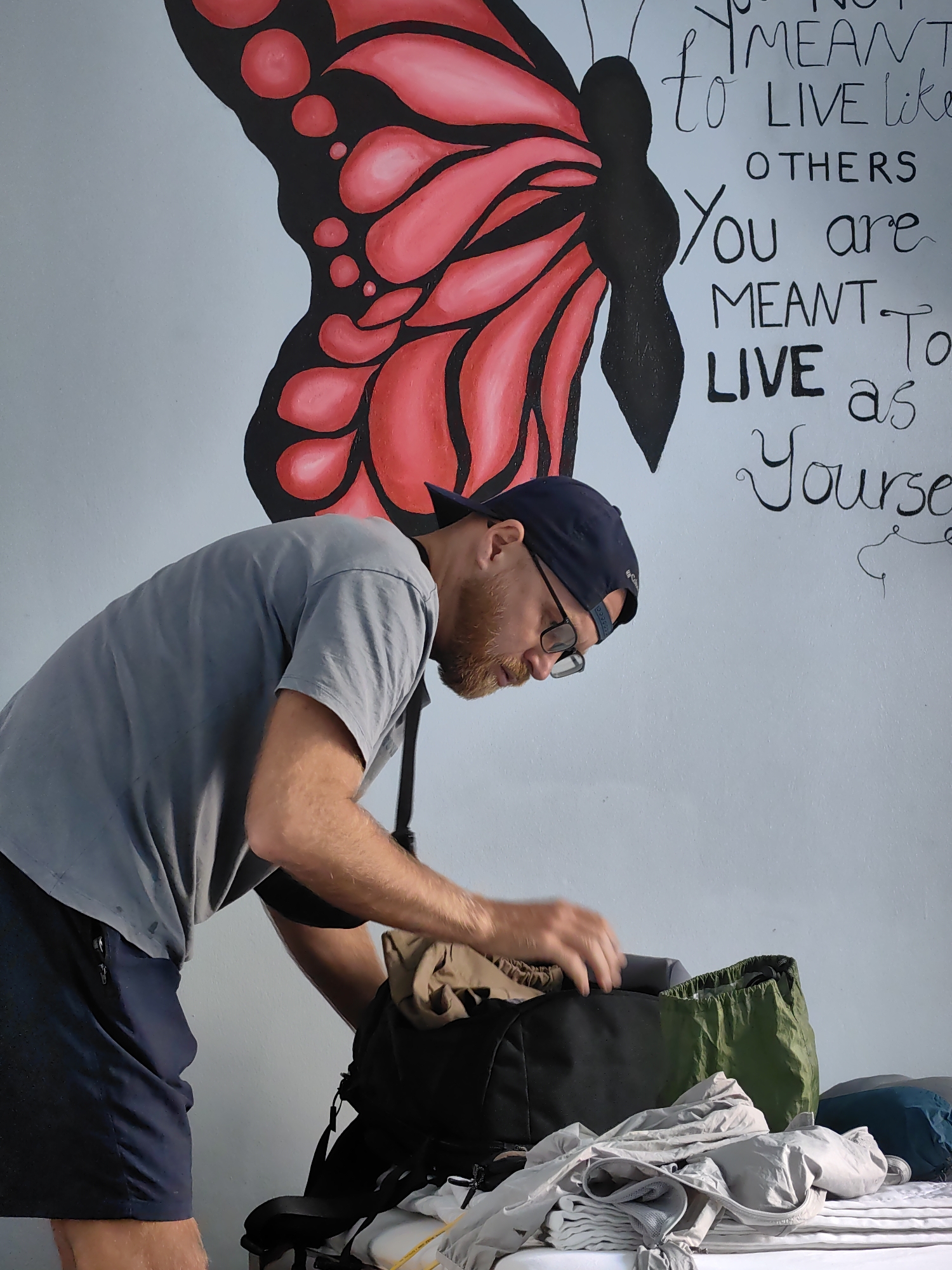 The image shows a man packing a black and green backpack in a room in Bai Chay Ward, Vietnam. The man is wearing a gray t-shirt, blue shorts, a blue baseball cap, and glasses. He is bending over the backpack, which is sitting on a white surface covered with various items of clothing. In the background, a wall is decorated with a painting of a red and black butterfly and handwritten text in black. The text reads, in part, "You are meant to LIVE as Yourself."