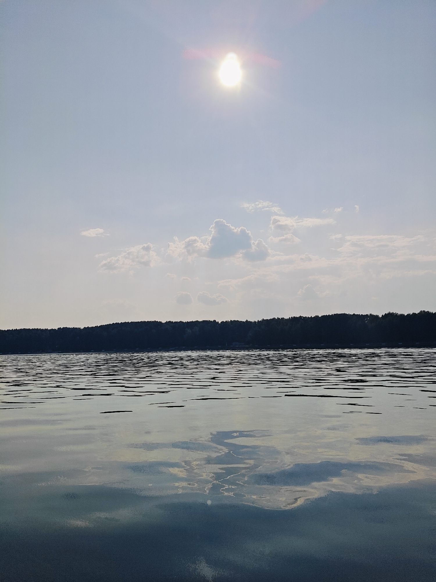 The image depicts a serene natural landscape with a body of water, a treeline, and a sky with clouds and the sun. The foreground is dominated by the calm surface of the water, reflecting the sky and the distant treeline. Small ripples distort the reflections, creating a textured pattern on the water's surface. In the middle ground, a dense, dark treeline stretches across the horizon, appearing as a silhouetted barrier against the sky. Above the treeline, the sky is a pale blue, with a few scattered white clouds. The sun is visible in the upper part of the frame, emitting a bright light with a lens flare. The scene appears to be a tranquil outdoor setting, likely a lake or a wide river. The image was taken from a low vantage point, close to the water's surface, emphasizing the expanse of the water and the sky. There are no people or man-made structures immediately visible. The overall impression is one of peace and natural beauty. This scene is consistent with the natural surroundings of Chaikovsky, Russia, a city situated on the Kama River, known for its picturesque landscapes.