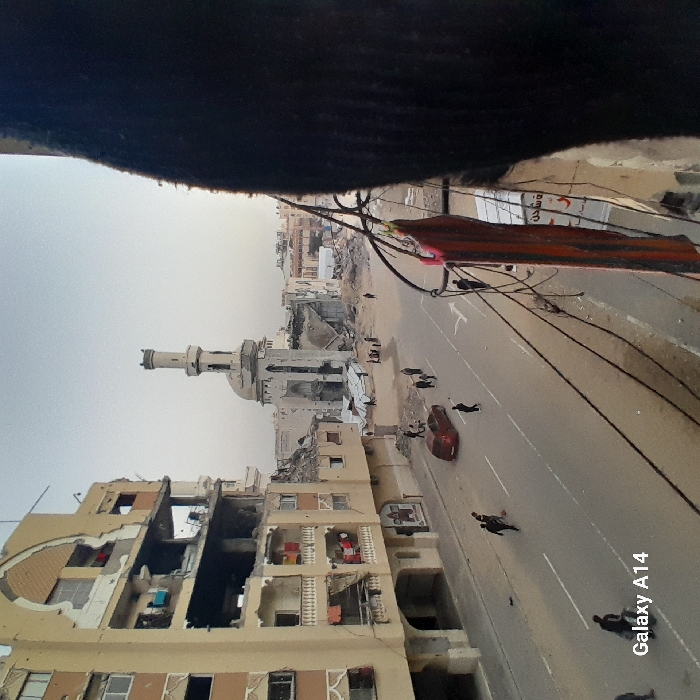 The image depicts a war-torn urban landscape. Buildings are heavily damaged, revealing exposed interiors. A mosque stands amidst the destruction, its minaret still intact. A road runs through the scene, with a red car and several people walking along it. Power lines crisscross overhead, adding to the sense of chaos. The sky is overcast, contributing to the somber atmosphere. A small inscription “Galaxy A14” is visible in the bottom right corner.