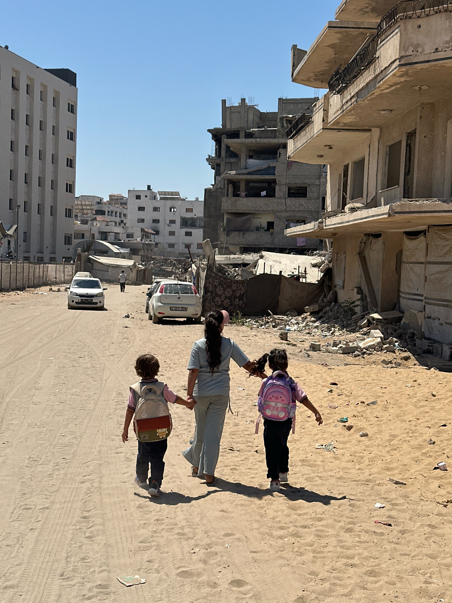 In Gaza City, Palestinian Territories, a woman walks with two children, a boy and a girl, down a dusty, unpaved road. The children, holding hands, wear backpacks, suggesting they are on their way to or from school. The boy has a beige and red backpack with a cartoon character, while the girl has a pink backpack with a cartoon pig and the number "08" on it. The woman is dressed in a light blue outfit.
The environment shows signs of conflict and destruction. To the right of the road, buildings are heavily damaged, with rubble scattered on the ground. Some structures appear to be partially collapsed or have gaping holes. To the left, a modern, multi-story white building stands intact, contrasting with the surrounding devastation. The road itself is sandy and appears to be a well-traveled path, with tire tracks visible. In the background, more damaged buildings and tents can be seen, indicative of a damaged urban landscape. Two white cars are parked on the road, and another person is walking in the distance. The sky is clear and blue, suggesting a sunny day.
