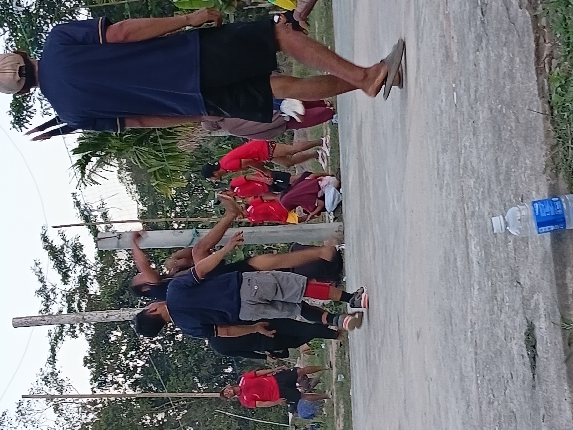 In Margo Rukun, Indonesia, a group of people are gathered outdoors on a concrete surface, likely a court or a yard. The main focus is on a volleyball game in progress, with several individuals actively participating. In the foreground, a person wearing a navy blue t-shirt and black shorts, with a tan baseball cap, is walking away from the camera, their feet in flip-flops. To their left, a person in a navy blue shirt and grey shorts is captured mid-air, appearing to be jumping or playing volleyball. Another person in a red shirt and black shorts is standing on the left, looking up. In the background, more individuals are visible, some in red shirts, and others in casual attire, with a backdrop of trees and foliage. A volleyball net and poles are present, confirming the sporting activity. A discarded plastic water bottle is on the ground in the lower right corner. The scene appears to be a casual, community sports event.