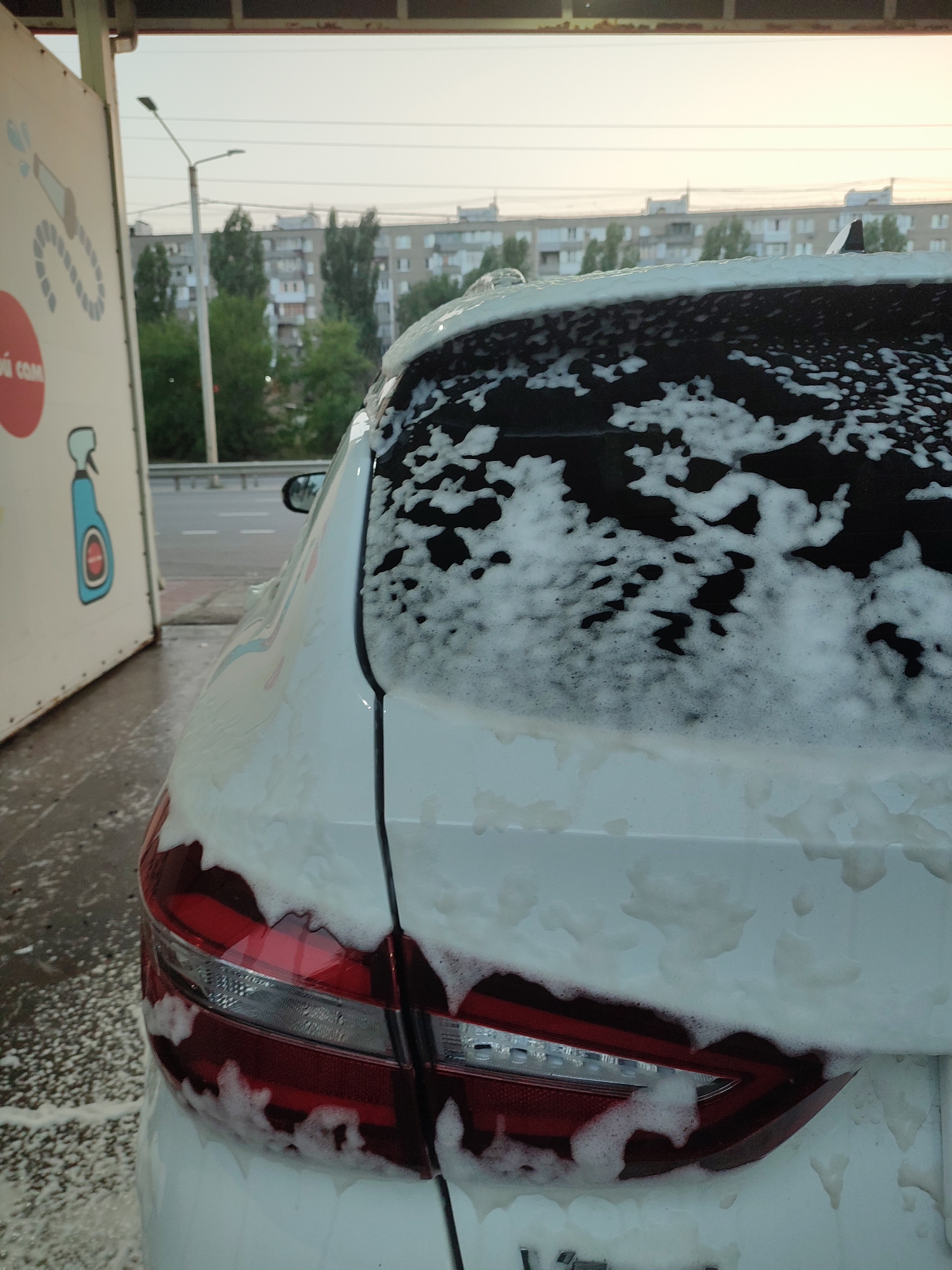A close-up shot shows the rear passenger side of a white car covered in white foam. The car is likely at a car wash, as indicated by the foamy residue on the ground and a white structure to the left with a graphic of a spray bottle and a hose. The car's taillight is visible on the lower left, and the rear window is heavily covered in foam, obscuring much of the view. In the background, across a road, are apartment buildings and trees, suggesting an urban setting. The sky is overcast, hinting at either dawn or dusk. This scene is consistent with an outdoor car wash facility in a residential area of Voronezh, Russia.