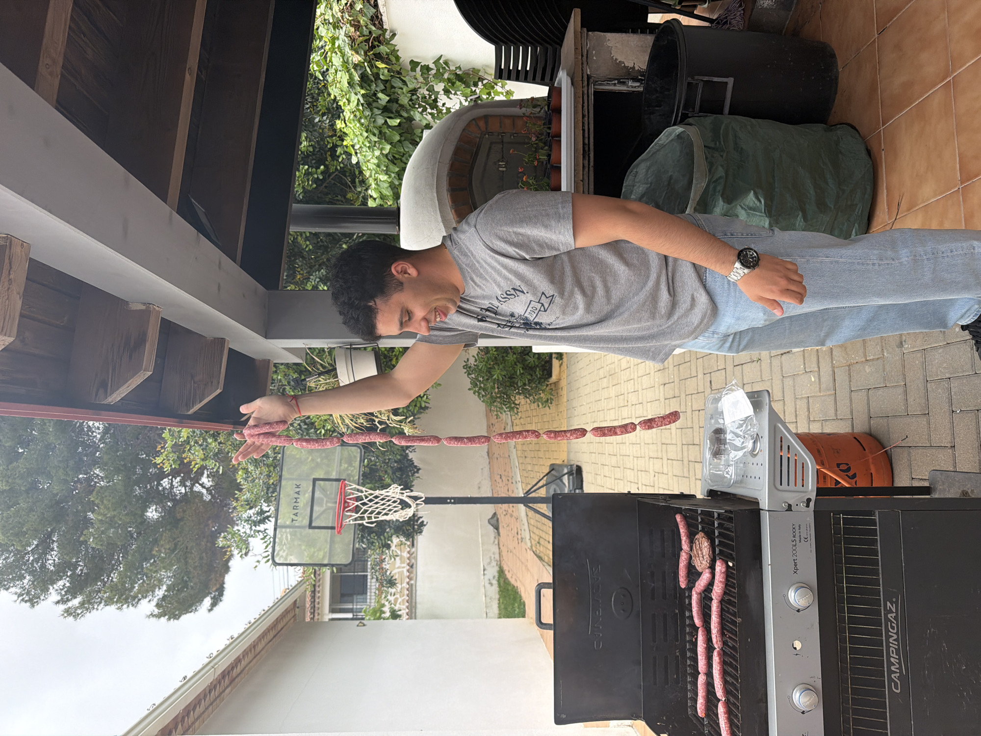 A man stands outdoors near a grill, holding a string of uncooked sausages in his extended hand. Some sausages are already cooking on the grill. A basketball hoop stands nearby in a yard surrounded by buildings and greenery. The location appears to be Montgó, Spain.