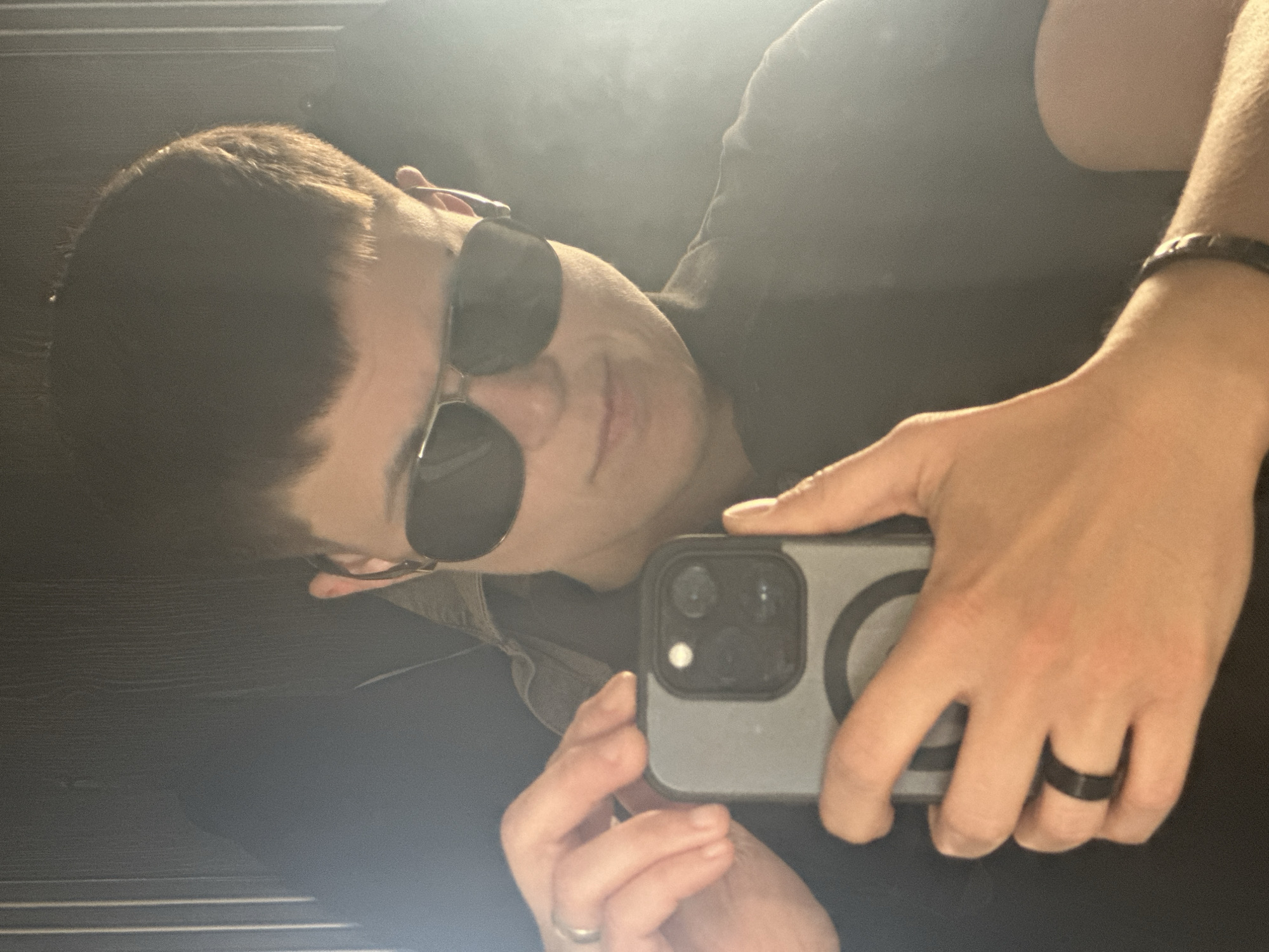 A person is taking a mirror selfie in what appears to be a dimly lit room. They are wearing dark sunglasses and a dark shirt. They are holding a smartphone in their hands, and are also wearing a ring on their left hand and a bracelet on their right wrist. The background is dark and out of focus. There are no discernible features to place this image in Uzhhorod, Ukraine.