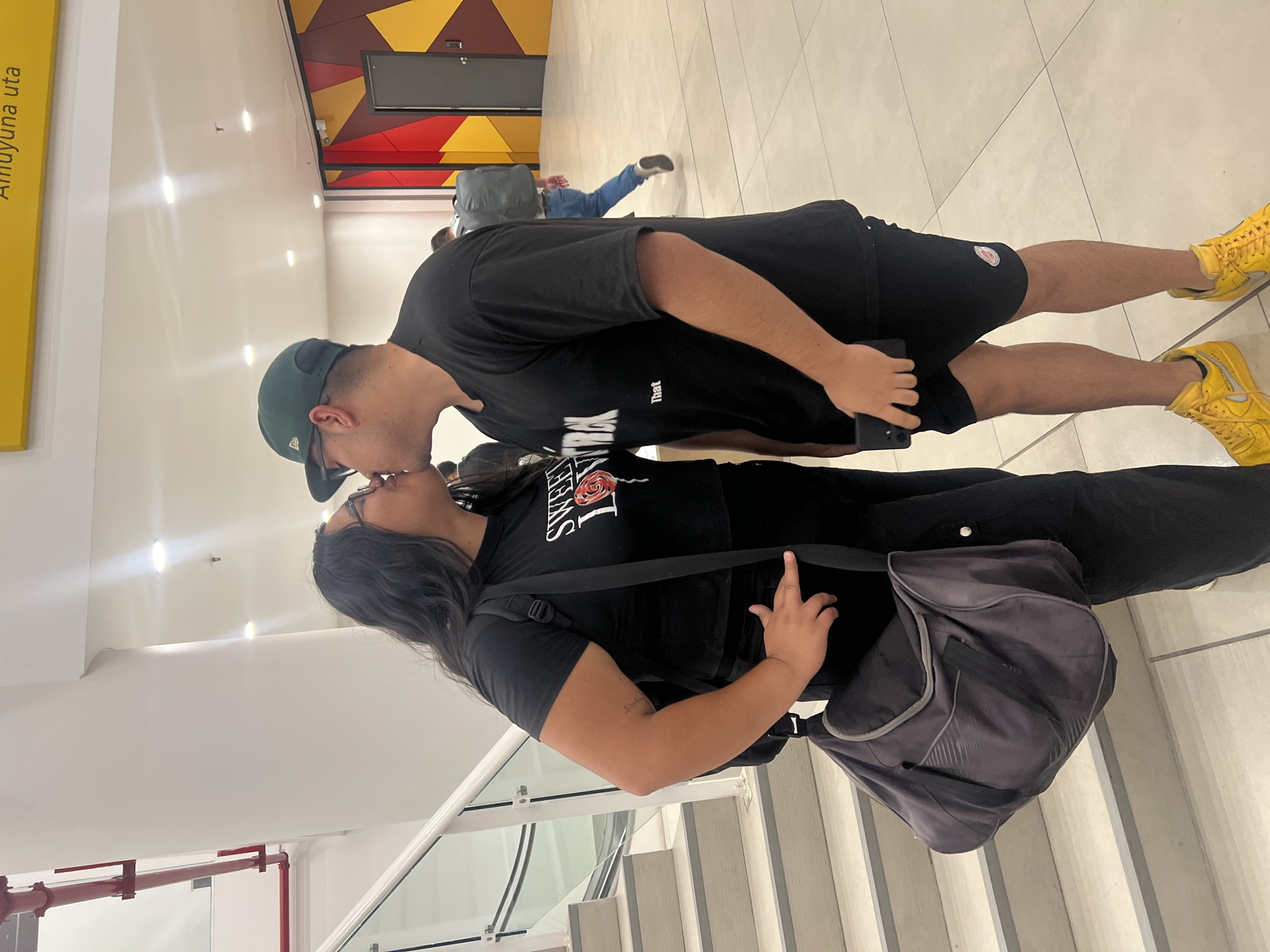 A couple is kissing in a bright, public space. The man is wearing a black t-shirt, black shorts, a green baseball cap, and yellow sneakers. He is holding a black smartphone in his right hand. The woman is wearing a black t-shirt with a rose graphic, black pants, and glasses. She is carrying a gray messenger bag. The background includes signage with "Amuyuna uta," a colorful geometric design, and escalators. Arica, Chile, is indicated by the signage. In the background, another individual can be seen walking in the distance.