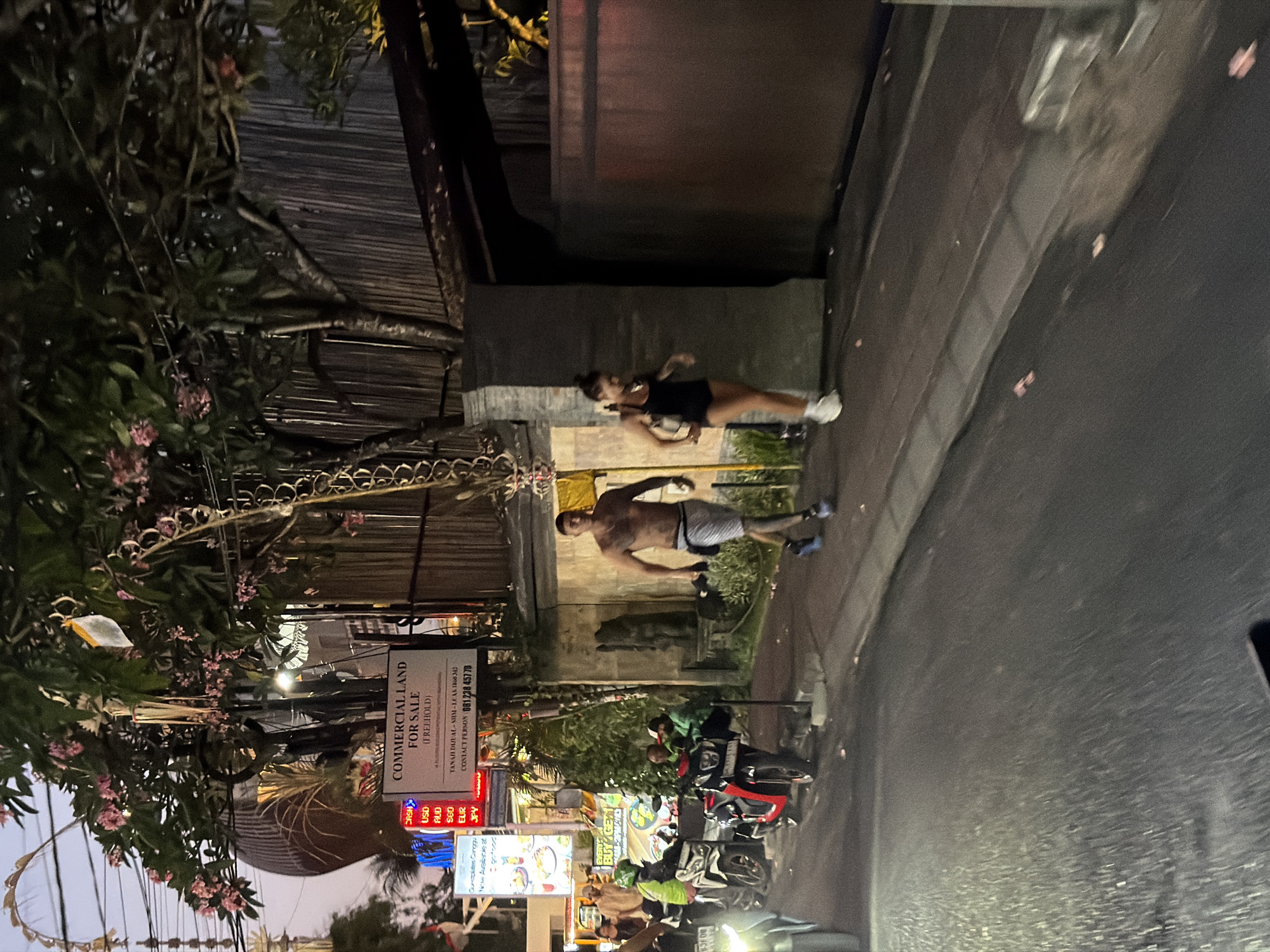 The image shows a street scene at dusk, with artificial lighting illuminating the surroundings. The perspective is from a low angle, possibly from a vehicle, looking up and across the street.
In the foreground, lush green foliage with pink flowers is visible on the left, partially obscuring a street sign that reads "COMMERCIAL LAND FOR SALE (FREEHOLD)". Below this, currency exchange rates for USD, AUD, SGD, EUR, and JPY are displayed. Several motorcycles are parked along the street, with some riders visible.
Further down the street, two people are seen standing. One, a woman in a black athletic top and shorts, is walking away from the camera. Another, a shirtless man with tattoos, is standing with his back to the camera, wearing light-colored shorts. They appear to be on a sidewalk or pathway next to the road.
The overall setting suggests an urban or semi-urban environment, likely in a commercial area, possibly in a tropical region given the vegetation. The time of day is indicated by the dim natural light and the presence of artificial lighting, suggesting either early morning or evening. The presence of currency exchange rates and a "For Sale" sign points to a commercial district.