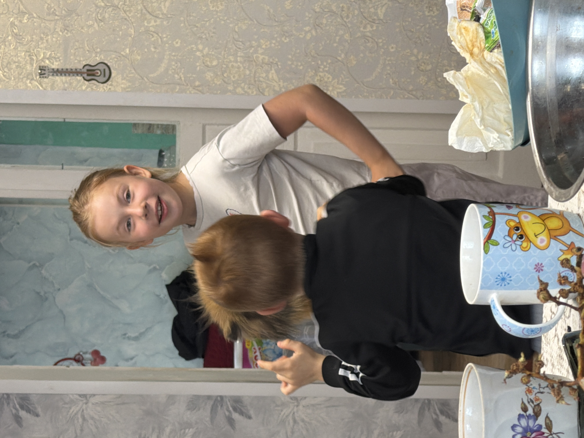 The image appears to be taken indoors, possibly in a home in Бронница, Russia, given the prompt. A young girl with blonde hair and blue eyes, wearing a light-colored shirt, looks at the camera with a smile. Beside her is a boy with reddish-brown hair wearing a black sweatshirt with white stripes on the sleeves. He seems to be looking at something near the bottom of the frame. In the background, there is wallpaper with a light floral pattern and a decorative guitar. In the foreground, a table holds several objects including mugs and a large metal bowl containing items wrapped in white paper. A mirror is positioned against a wall displaying images of items on a shelf or table.