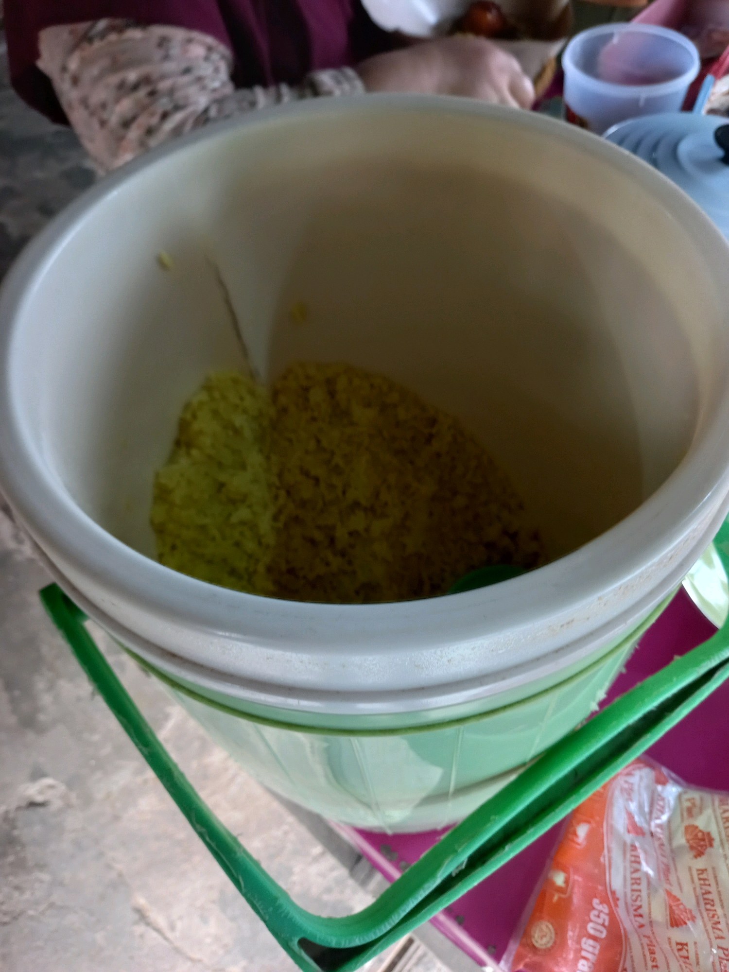 A close-up shot shows a large, light-colored plastic tub containing a yellowish, granular substance. The tub has a light green rim and a green plastic handle. In the background, a person's hands are visible, along with other objects like a blue pot lid and a clear plastic cup containing a pink liquid. A package labeled "KHARISMA Plast" and "350 gram" is partially visible in the lower right corner. The scene appears to be indoors, likely in a residential setting in Samarinda, Indonesia, possibly during food preparation or storage.