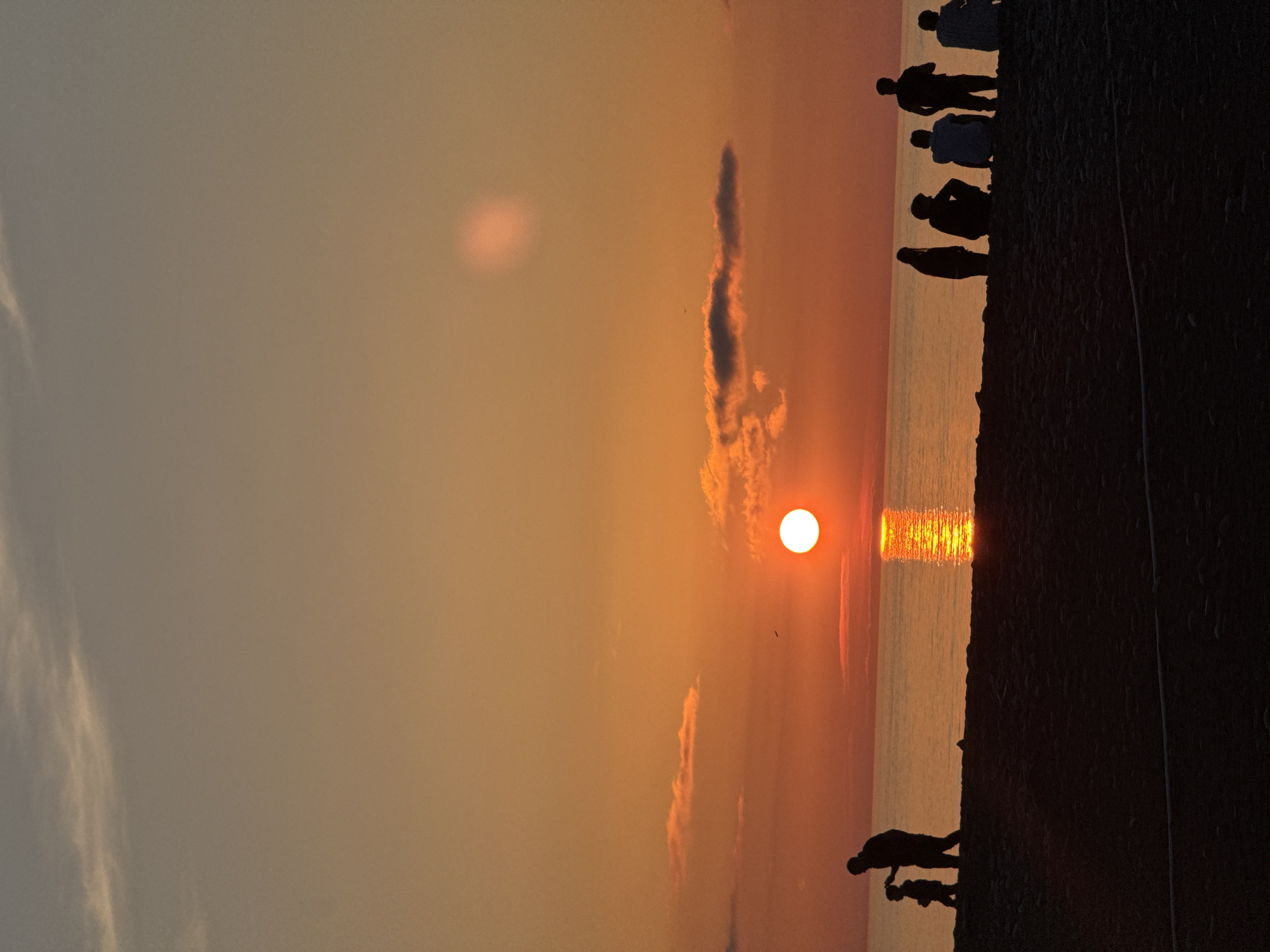 A photograph captures a sunset scene at a seaside location, likely in Batumi, Georgia, based on similar images. The image is oriented vertically. A bright, circular sun is positioned near the horizon, reflected on the surface of the water, creating a path of light. Silhouetted figures are visible along the shoreline. The sky ranges in color from orange near the horizon to a lighter hue higher up. Some cloud formations are present, partially obscuring the sun. The foreground shows a dark, textured area, possibly a seawall or embankment. Silhouetted people are either walking along the beach or standing on the edge of the shore. At the bottom of the frame, a man appears to be crouched down, possibly taking a picture or interacting with a child. The figures are small and indistinct due to the backlighting of the sunset.