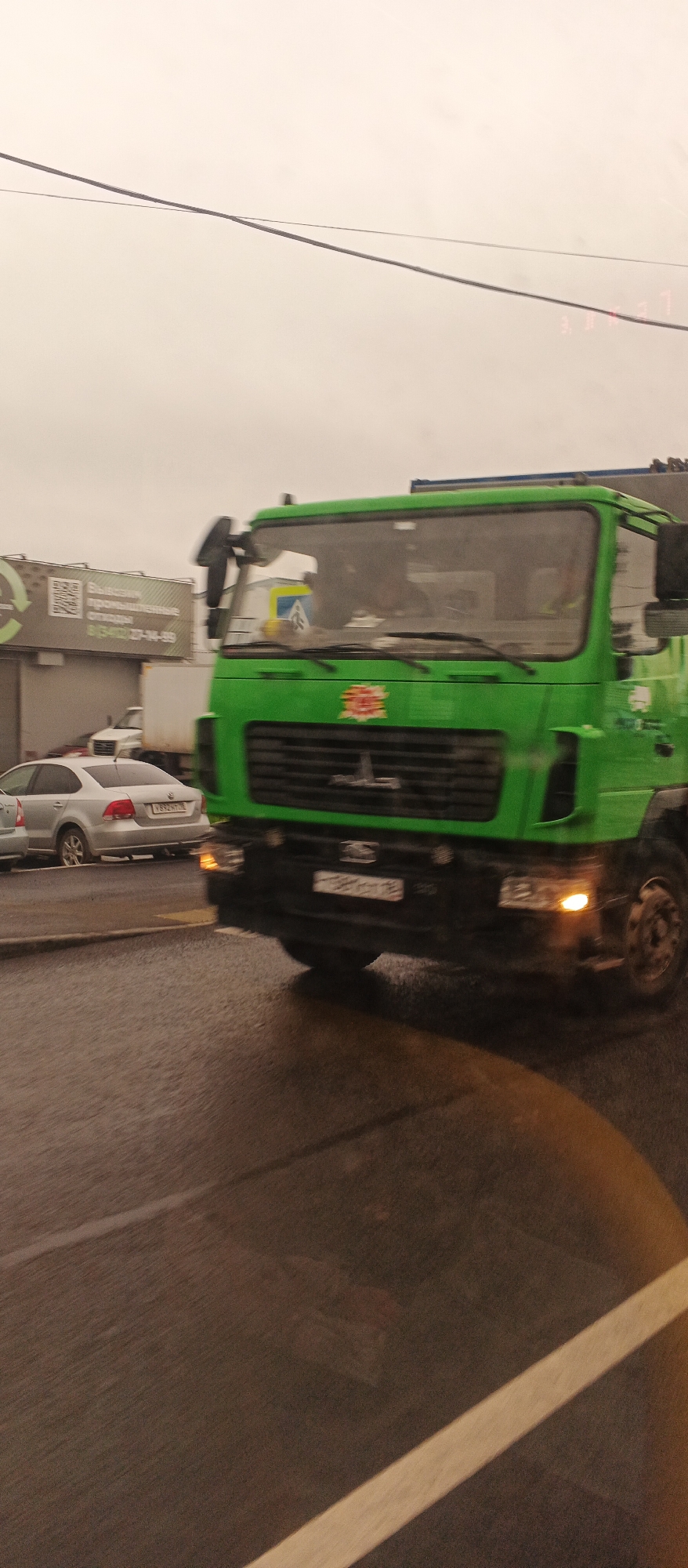 An outdoor, eye-level, slightly blurry, medium shot shows a green truck driving on a wet, paved road in Izhevsk, Russia. The truck has a large grille, a prominent logo on the front, and a yellow and blue sign inside the windshield. The sky is overcast and grey. To the left of the truck, a silver sedan is parked on the side of the road. In the background, a billboard with a QR code and Russian text is visible. The scene appears to be on a cloudy day, possibly after rain, as the road surface is reflective.