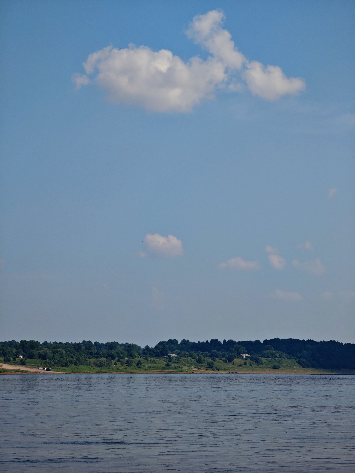 The image depicts a wide, calm river flowing in front of a densely forested embankment under a bright blue sky with scattered white clouds. On the embankment, which appears to be the outskirts of Ust'-Kem', Russia, there are a few scattered buildings and some vehicles parked on a dirt track. The water surface is rippled, reflecting the light of the sky. The overall scene is serene and natural, with a hint of human presence on the distant shore.