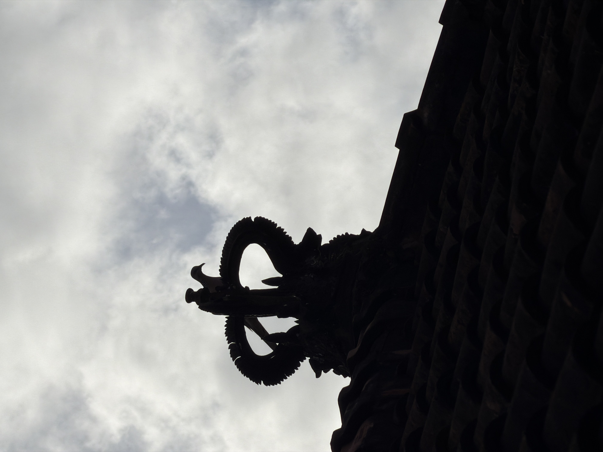The image shows a close-up, silhouetted view of a Balinese dragon statue projecting from the corner of a dark building or structure, set against a cloudy sky. The statue is intricate with curled horns and detailed features. The building appears to have a textured surface, potentially made of dark wood or stone. The location is likely in North Kuta, Indonesia, considering the Balinese architectural style of the dragon statue. The scene is captured from a low angle, emphasizing the statue's placement on the building's edge.