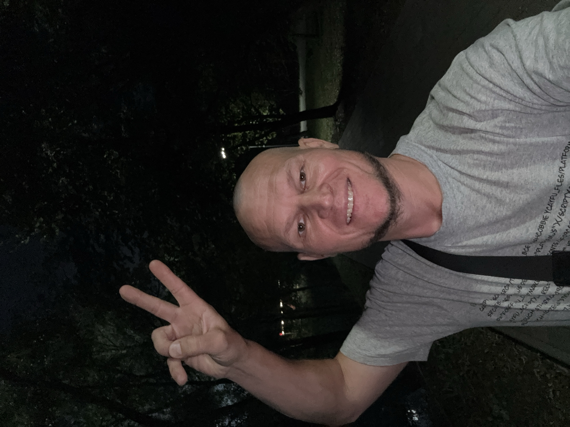 A middle-aged bald man with a goatee and a grey t-shirt stands outdoors at night, making a peace sign with his right hand. He has a black strap across his chest. The background shows dark trees and a lamppost. The t-shirt has text printed on it that appears to be code. The location is not identifiable as Oskemen, Kazakhstan.