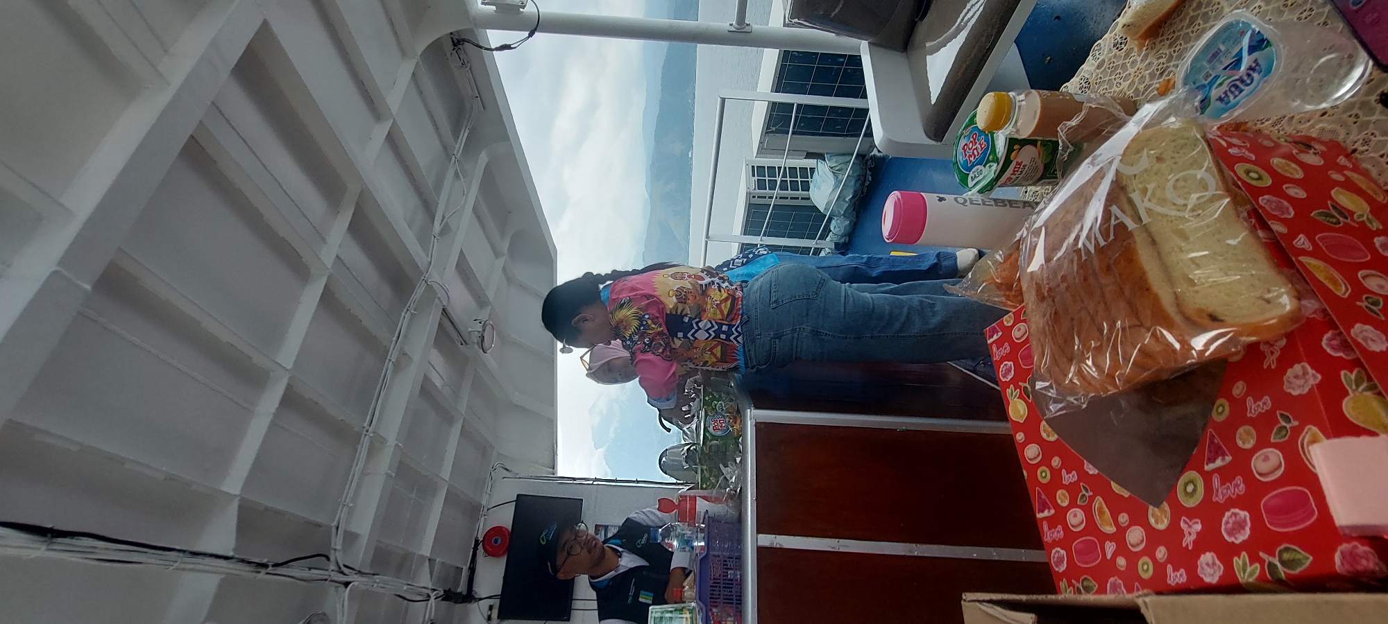 The image depicts an indoor scene on what appears to be a ferry or boat. The ceiling is made of white panels with visible wires running across. In the background, a body of water and distant hills are visible through a window or opening.
There are three individuals in the foreground. On the left, a person wearing a dark cap, glasses, and a blue vest with logos is standing behind a counter, looking down. To the right of this person, two women are interacting. One woman, wearing a pink headscarf and a colorful patterned shirt, is looking towards the left. The other woman, wearing a pink headscarf and a vibrant, patterned top with blue jeans, is facing away from the camera, and appears to be handing something or interacting with items on the counter.
On the counter, there are various objects, including plastic cups, bottles of water, and containers of food. A large loaf of bread, partially wrapped in plastic, is prominently displayed on a red, decorated box in the lower foreground. Other food items, including a cup labeled "POP MIE," are also visible on the counter. The flooring is a dark blue color. The overall impression is of a casual gathering or service area on a vessel. The specific location in the city of Hobong, Indonesia, cannot be determined from the visual information in the image.