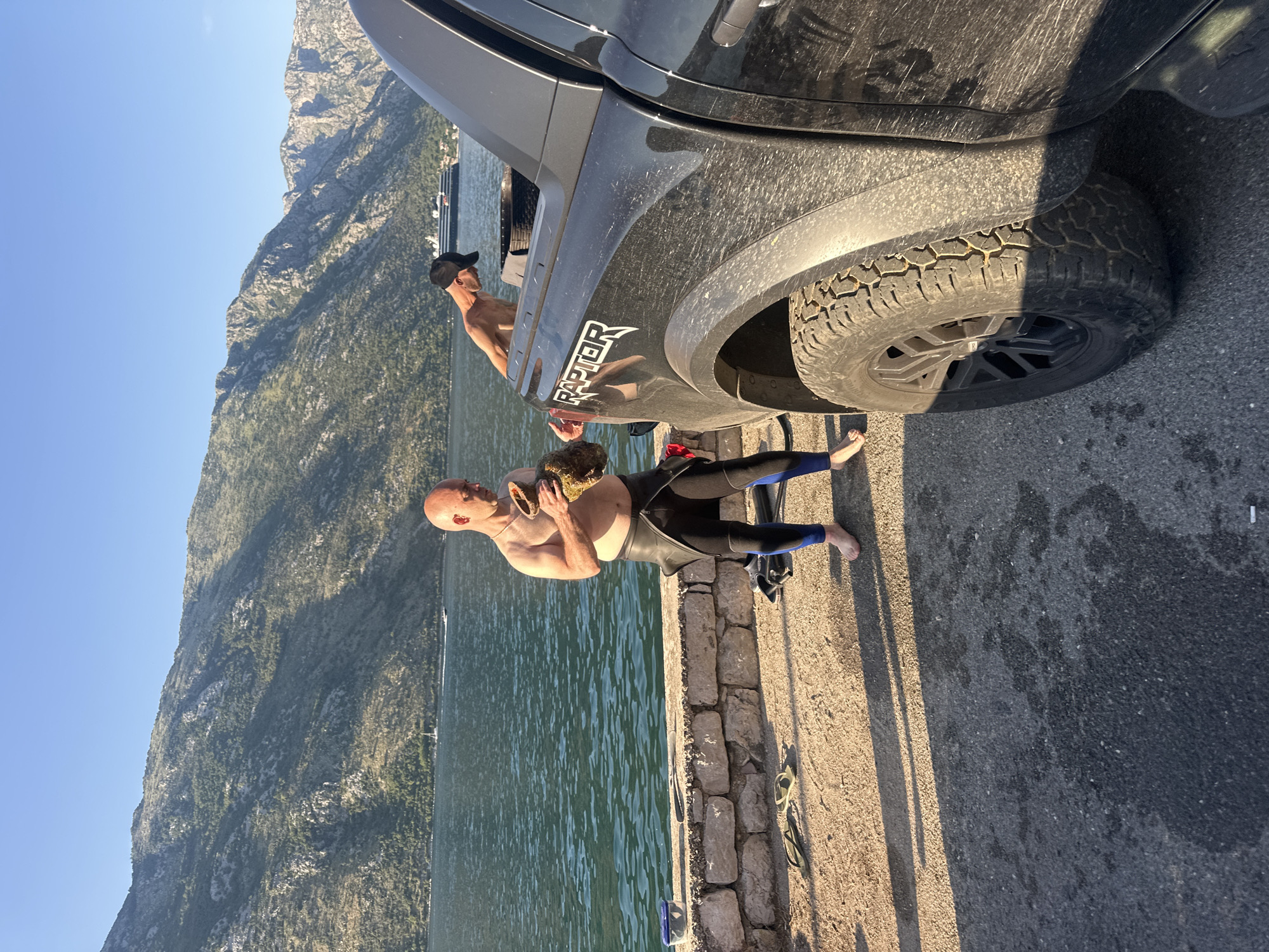 The image depicts a coastal scene in Prčanj, Montenegro, featuring two men and a dark pickup truck by a body of water with mountains in the background under a clear sky.
In the left-center, a bald, shirtless Caucasian male with a light tan is standing barefoot on a paved surface. He is wearing the lower half of a black and blue wetsuit, with the top half pulled down to his waist. He is holding two irregular, organic-looking brown objects in his hands, appearing to examine them. One object, larger, is in his left hand, and a smaller one in his right. His gaze is directed downwards at these objects.
To the right, partially obscured by a dark Ford Raptor pickup truck, a second leaner Caucasian male, wearing a black baseball cap and bare-chested, is bent over. He appears to be interacting with the bed of the truck, possibly retrieving or stowing items. The truck is dark-colored, visibly dusty, and features "RAPTOR" branding on its side. A robust tire with an aggressive tread pattern is visible on the front passenger side.
The ground consists of a paved surface (asphalt or concrete) in the foreground, transitioning to a stone wall or embankment directly adjacent to the water. A pair of yellow-green flip-flops and a blue plastic container with a clear lid are resting on the paved ground near the wall.
The body of water in the background is calm with visible ripples, reflecting the sunlight. Beyond the water, steep, rocky mountains covered with sparse vegetation rise prominently. A large cruise ship is visible in the mid-distance on the water, positioned to the left of the second man and the truck. Smaller boats, including a sailboat, are also discernible further in the distance. The lighting suggests either morning or late afternoon, with strong, directional sunlight creating distinct shadows on the ground.