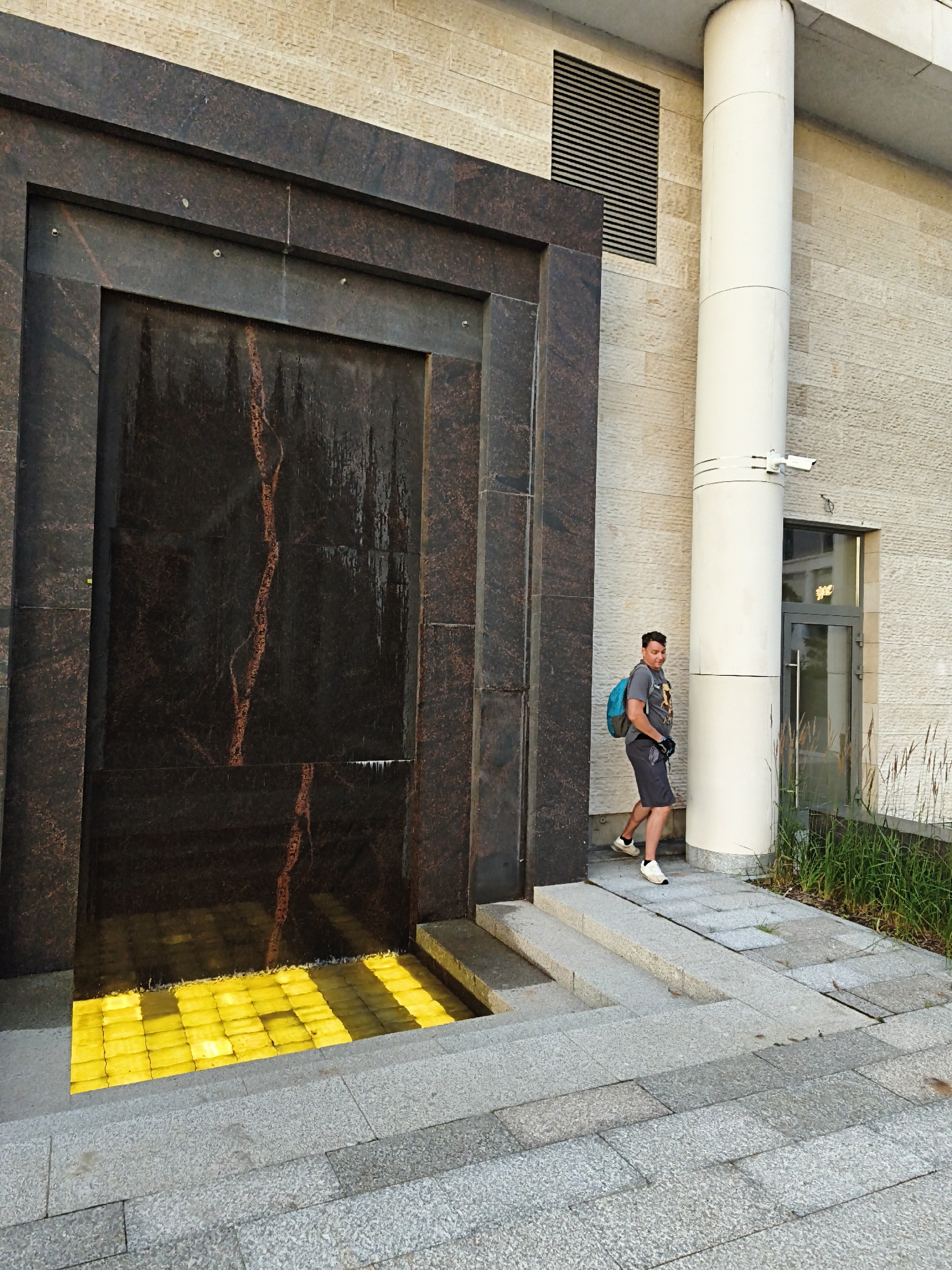 An outdoor shot captures a man with dark hair, wearing a grey t-shirt, dark shorts, black gloves, and white sneakers, walking towards a building. He is also wearing a bright blue backpack. The man is positioned to the right of the frame, near a large white cylindrical pillar. To the left of the pillar, there is a dark granite facade with a waterfall feature. The waterfall is a dark rectangular panel with a reddish vein running through it. Below the waterfall is a pool illuminated with bright yellow lights, creating a striking contrast. The ground and stairs are paved with granite tiles. The building's facade is made of textured, light-colored stone. A ventilation grate is visible on the wall above and to the left of the waterfall. A security camera is mounted on the white pillar. The scene appears to be an urban setting, possibly a modern building's entrance or a public space.