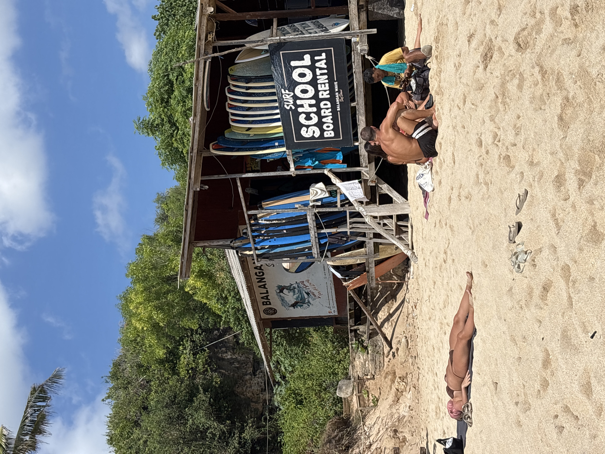 This image depicts a sunny beach scene at Balangan Beach in Kuta Selatan, Indonesia, featuring a surf school and individuals relaxing on the sand. In the center-right, a rustic, open-sided wooden shack with a corrugated metal roof serves as a "BALANGAN SURF" and "SURF SCHOOL BOARD RENTAL" establishment, indicated by prominent black and white signage. Numerous surfboards, predominantly blue and white, are vertically stacked within and around the structure. Lush green tropical vegetation covers a hillside behind the shack, under a clear blue sky with scattered white clouds.
In the lower-left foreground, a woman with light-colored hair and tanned skin lies supine on a brown towel on the golden-brown sand, wearing a dark brown bikini and appearing to be sunbathing. A black bag and a pair of light-colored sandals are positioned near her head. Closer to the shack, three individuals are seated on the sand. One man, with a muscular back and dark hair, wears dark swim shorts with white stripes and faces towards the shack. A woman with dark hair sits next to him, partially obscured. To their right, a man in a yellow t-shirt and a light blue towel draped over his shoulders is looking down at an object in his lap, with a black bag beside him. Additional pairs of flip-flops are scattered on the sand in the mid-ground. The scene is illuminated by direct sunlight, casting distinct shadows.