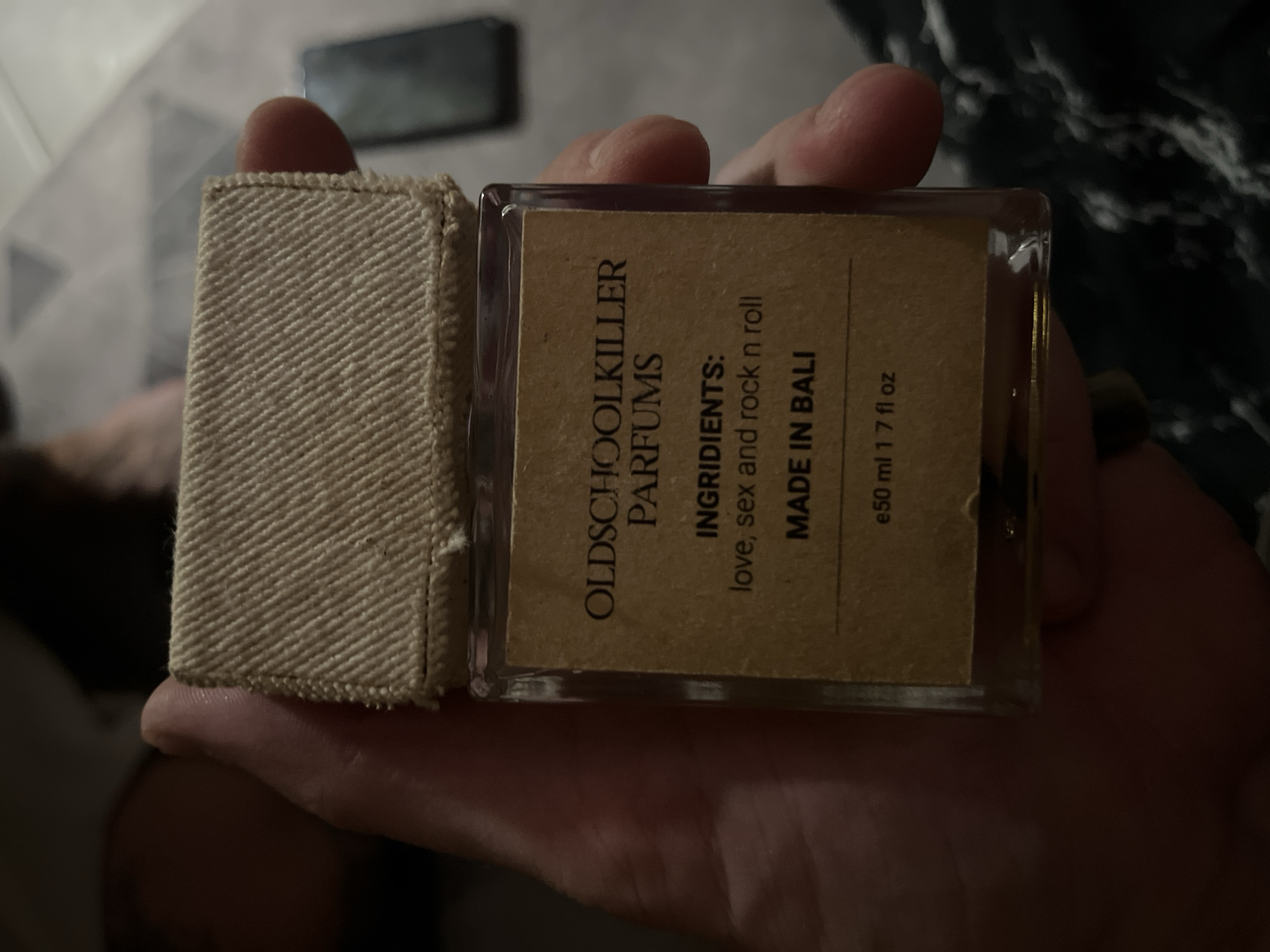 A person is holding a small glass perfume bottle with a fabric cap. The bottle has a brown label with black text. The text reads "OLDSCHOOLKILLER PARFUMS", "INGREDIENTS: love, sex and rock n roll", "MADE IN BALI", and "e50 ml 1.7 fl oz". The background is dark and out of focus, with what appears to be a phone or tablet on the right side. The location is not specified in the image.
