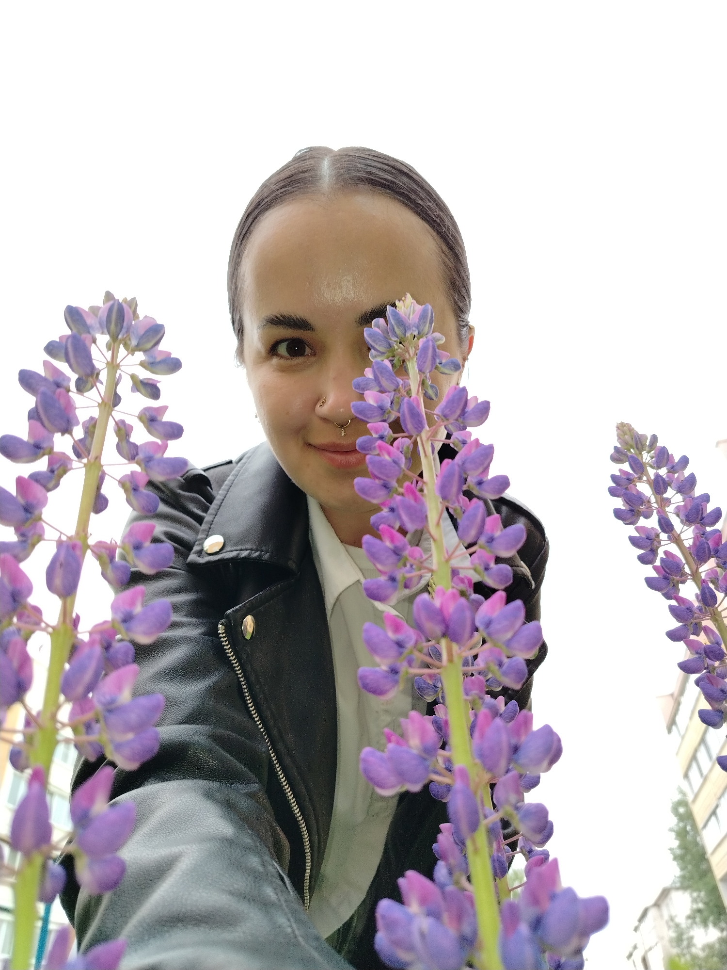 The image displays a woman from a low-angle perspective, positioned behind and between several stalks of purple and pink lupine flowers. She has dark hair pulled back, brown eyes, and a subtle smile directed at the camera. She is dressed in a black leather jacket over a white collared shirt. Her right nostril features a small stud piercing, and her left nostril has a ring with a small decorative element. The foreground is occupied by the in-focus lupines. The background consists primarily of a bright, overexposed white sky, with portions of light-colored, multi-story residential buildings and sparse green foliage visible in the distant right. The scene is situated in Noviy, Russia.