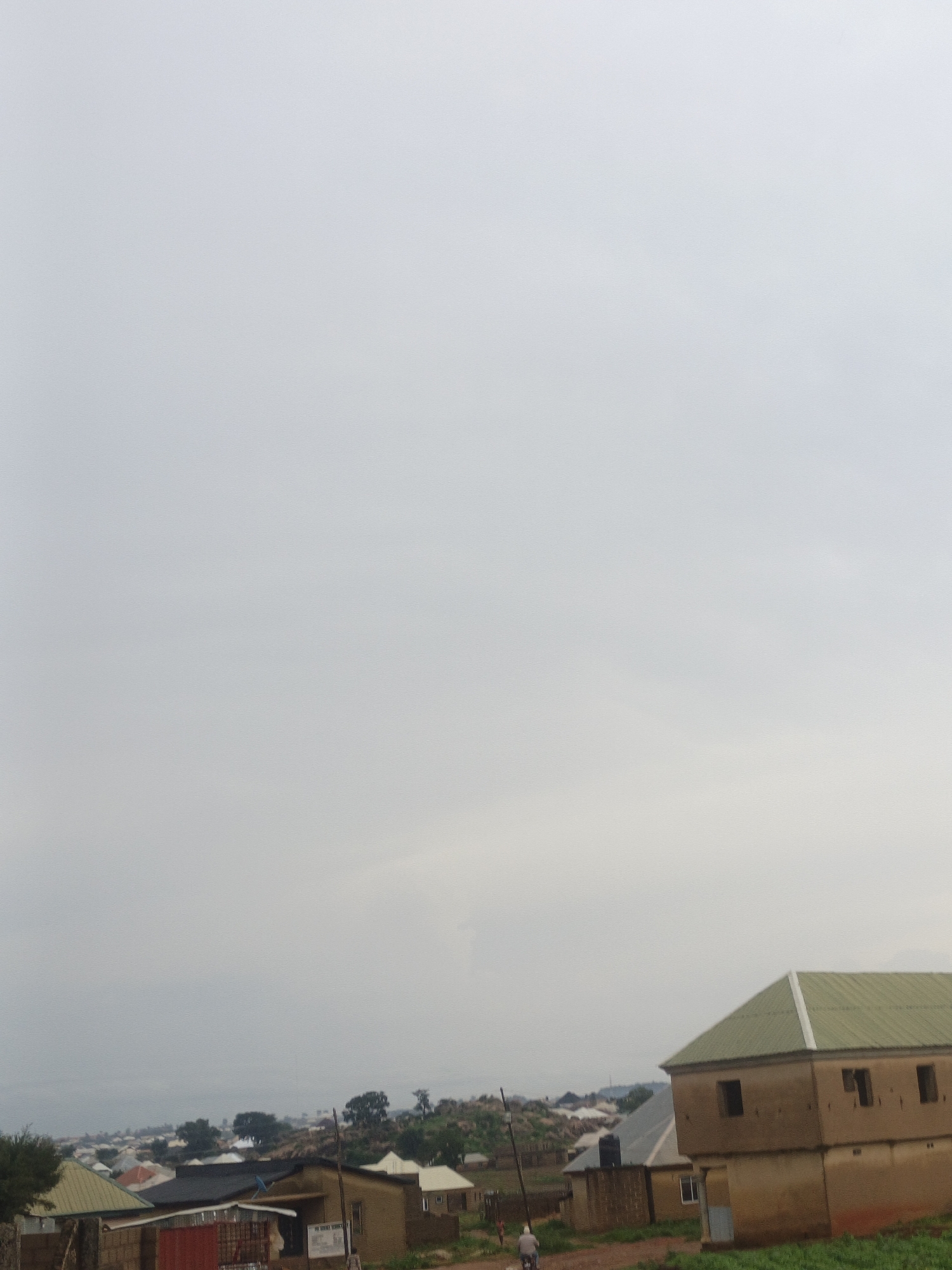 The image depicts a rural or semi-rural landscape, likely in the city of Rafin Bauna, Nigeria, under an overcast sky. In the foreground, a light brown, two-story building with a green corrugated metal roof and visible windows is prominent. To the left of this building, another structure with a dark roof is partially visible, along with a red metal gate or fence. In the middle ground and background, a collection of houses with various roof types, including some with corrugated metal, are spread across rolling hills dotted with trees. A single person is visible on a motorcycle in the lower right quadrant of the image, moving away from the viewer. The overall impression is one of a developing community with a mix of traditional and modern housing.
