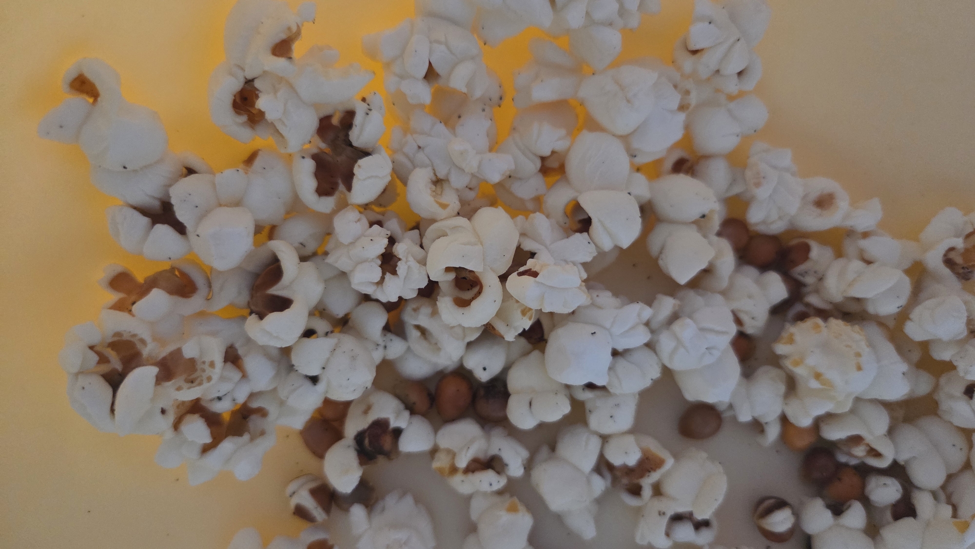 A close-up, overhead shot displays a pile of freshly popped popcorn and several unpopped kernels against a smooth, light yellow background. The majority of the frame is filled with white, irregularly shaped pieces of popped popcorn, many of which still have visible brown or tan remnants of the kernel attached. Scattered among the popped kernels are numerous small, round, brown unpopped popcorn kernels. The lighting is soft and even, highlighting the varied textures of the popcorn. No human figures or activities are depicted, presenting a static scene. There is no visible text. The setting appears to be an indoor surface, possibly a bowl or counter. The image provides no specific cues regarding time of day or weather, nor any direct visual confirmation of the given location context of Chaikovsky, Russia.